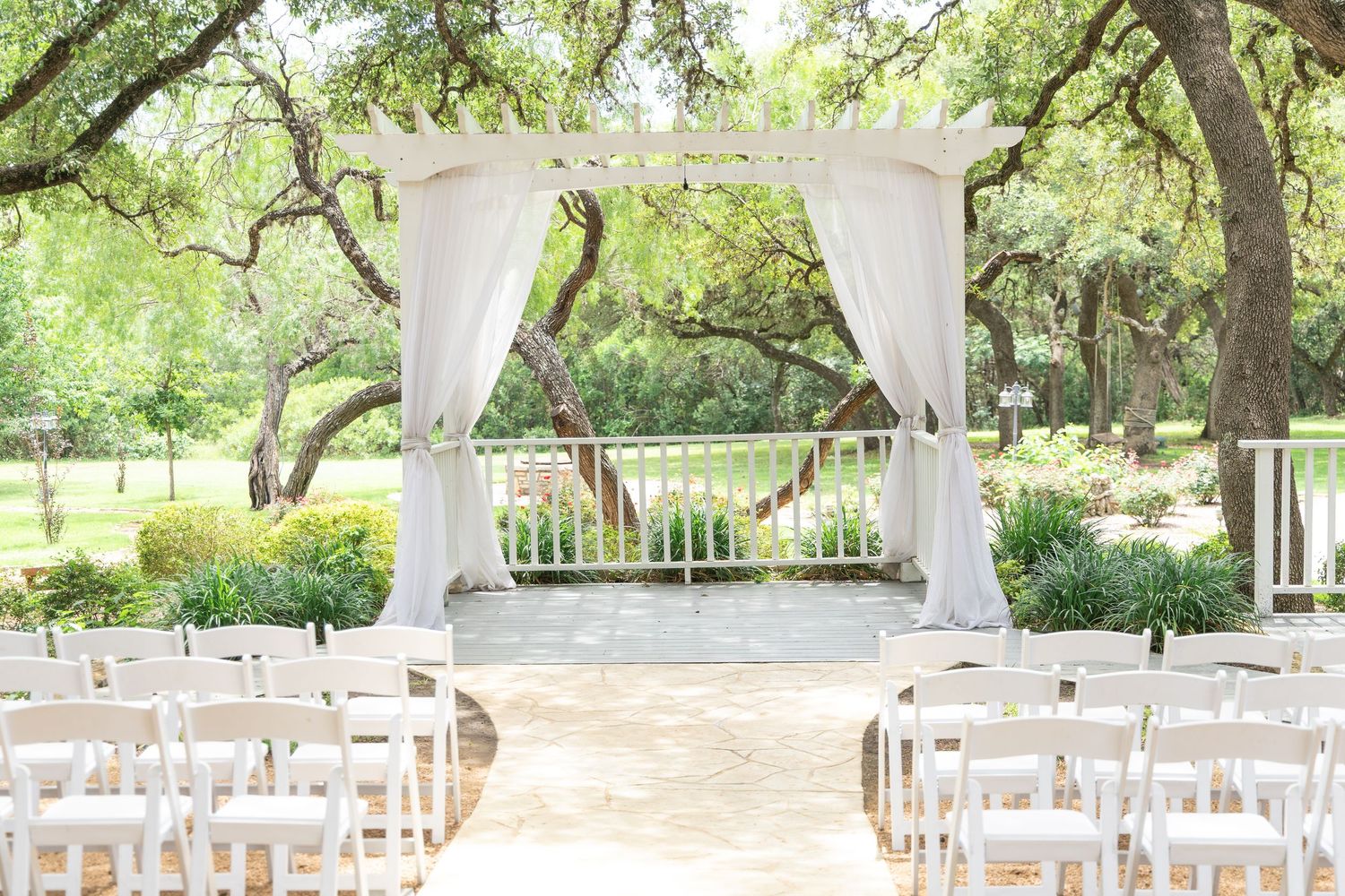 Elegant outdoor wedding ceremony venue with white draped arbor, white folding chairs, and lush green trees.