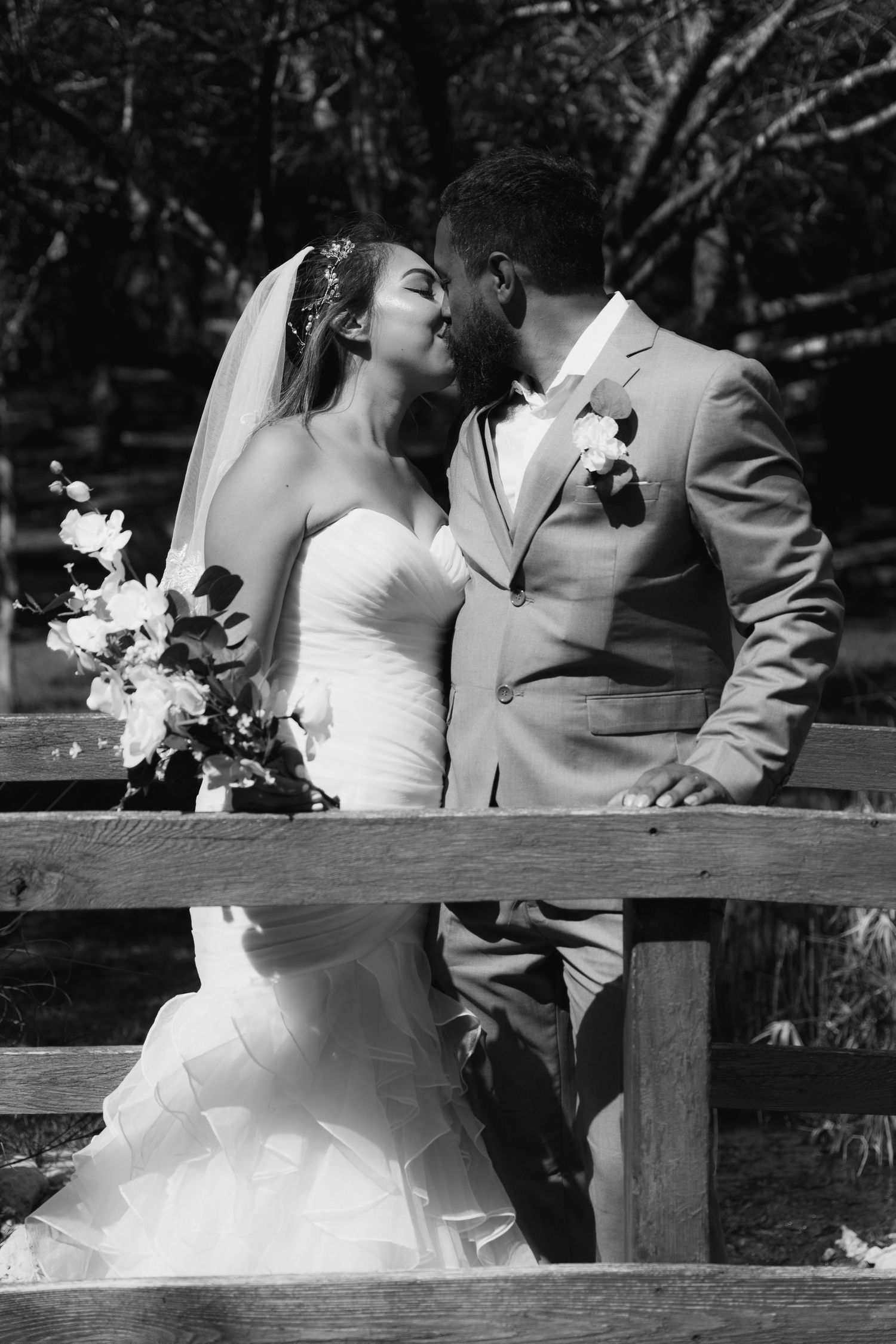 Black and white photo of bride and groom sharing a kiss near a wooden fence outdoors.