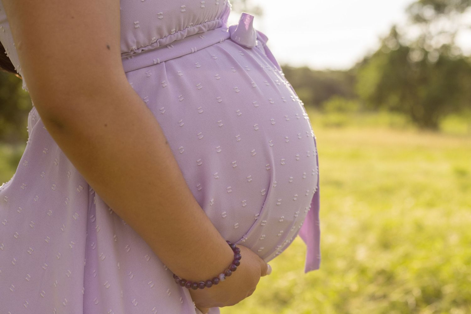 Close-up of pregnant woman in lavender polka dot dress cradling baby bump outdoors.