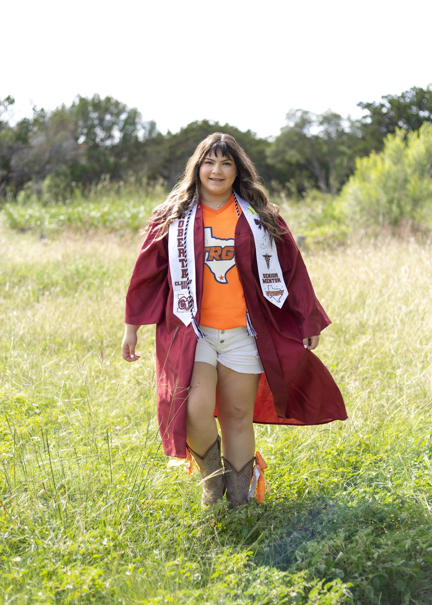 Graduate in maroon cap and gown walking through green field wearing orange Texas shirt and cowboy boots.