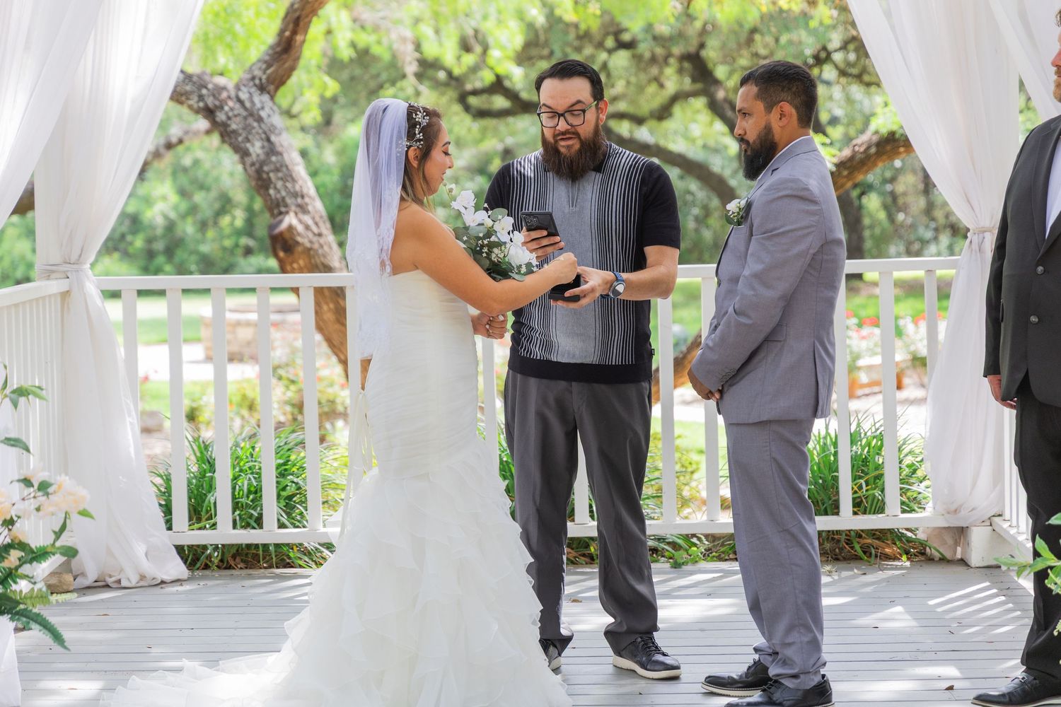 Wedding officiant places ring on brides hand during outdoor ceremony under white draped pergola.