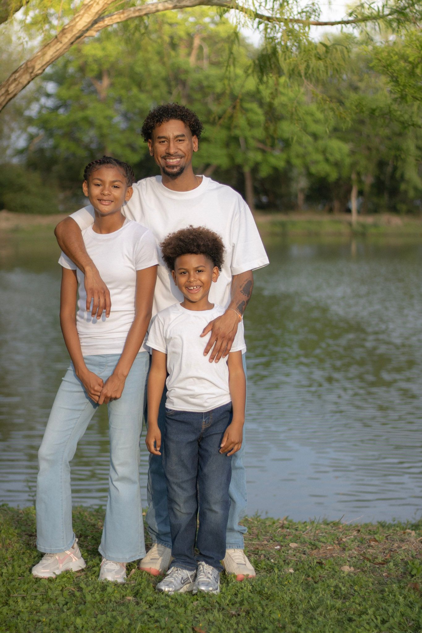 Father posing with his two children in white tees and jeans beside a calm park pond.