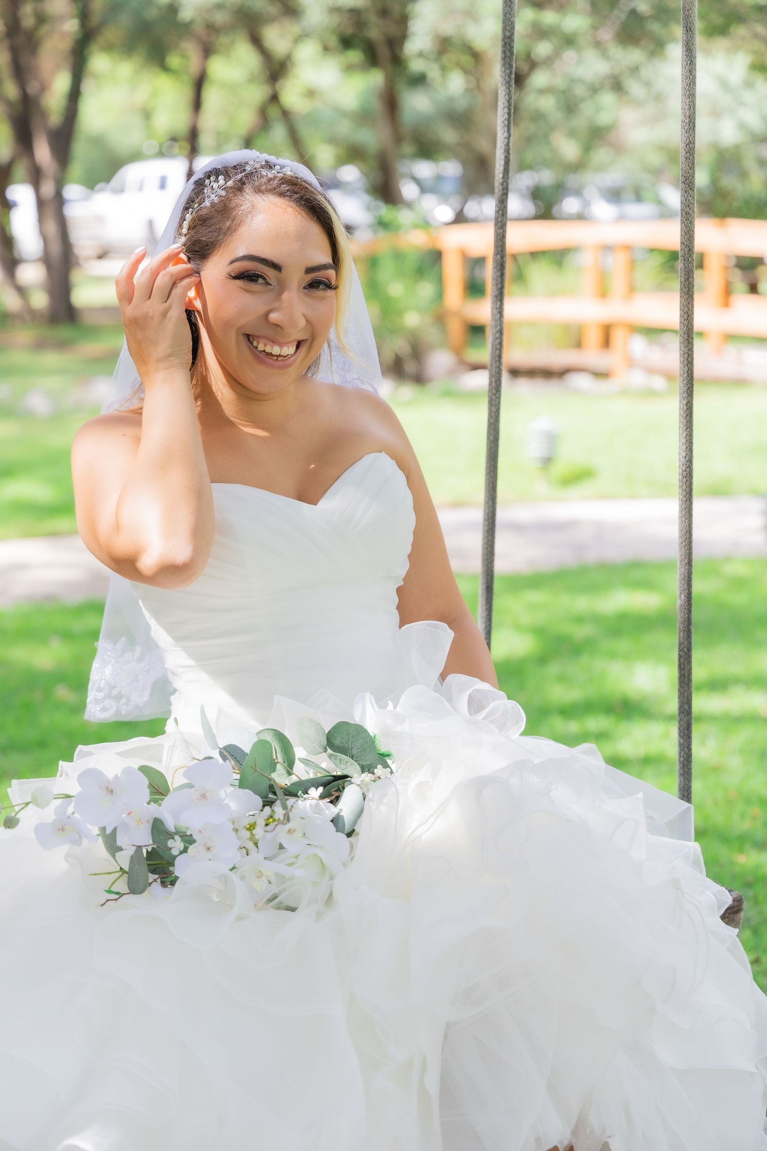 Smiling bride in white strapless gown sitting on swing holding white floral bouquet outdoors.