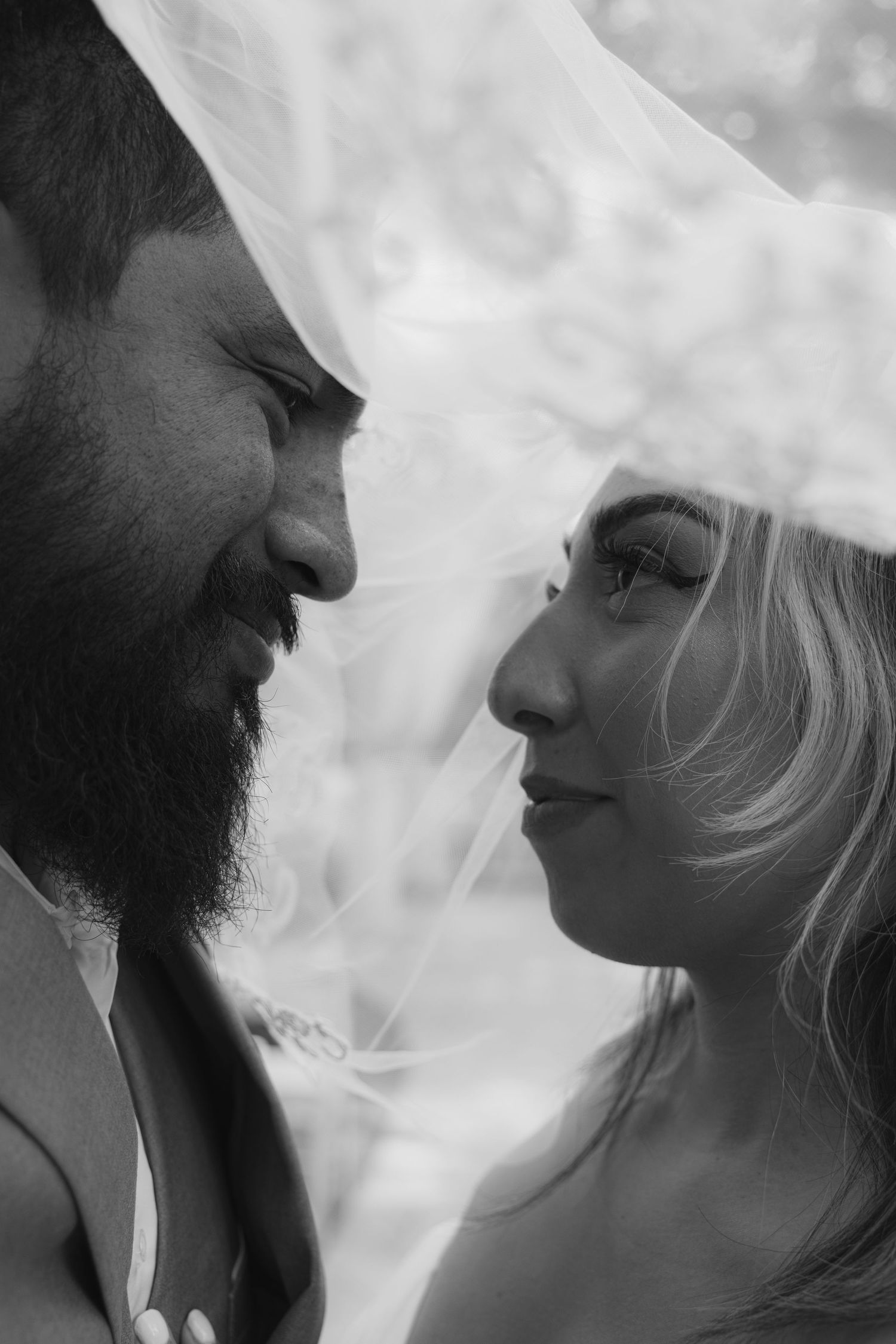 Black and white close-up of bearded groom and blonde bride gazing at each other beneath a bridal veil.