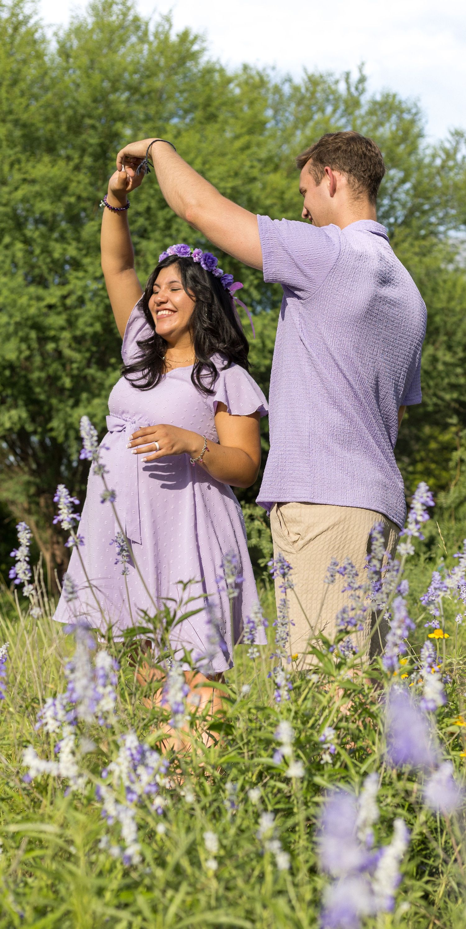 Happy pregnant woman in lavender dress dancing with partner in blooming wildflower meadow.