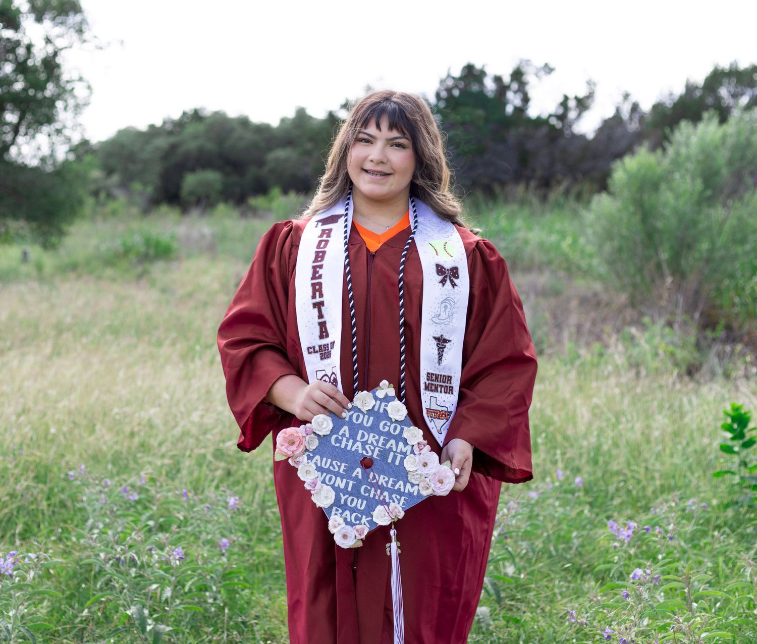 Young woman in maroon graduation gown and cap holding decorated mortarboard in green meadow.