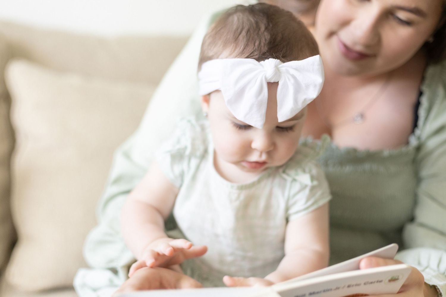 Close-up of baby girl with white bow headband reaching toward board book held by mother.