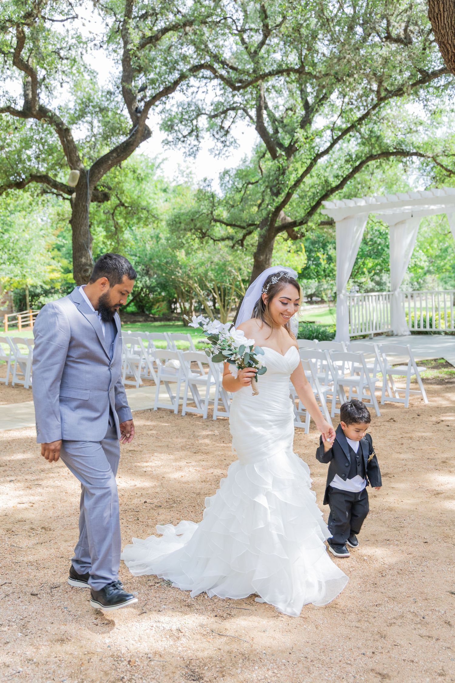 Bride in mermaid wedding gown walks with groom in gray suit and young boy in tuxedo at outdoor ceremony.