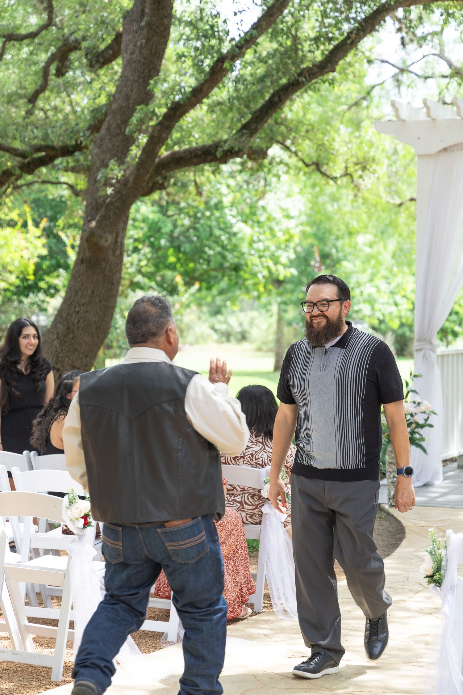 Man in black polo shirt smiling and greeting guest at outdoor wedding ceremony under large oak trees.