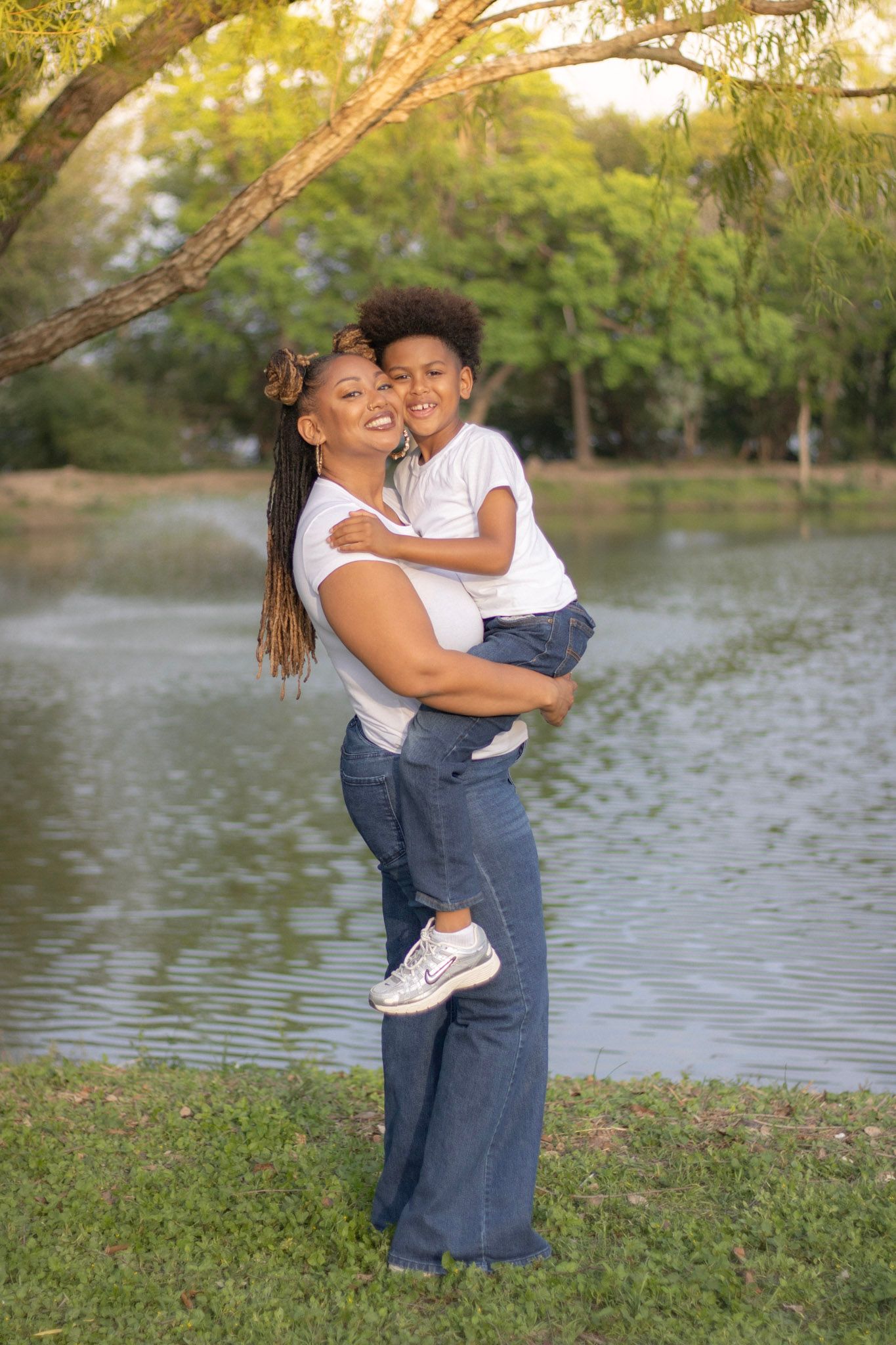 Smiling mother holding her curly-haired son in her arms beside a peaceful park pond.