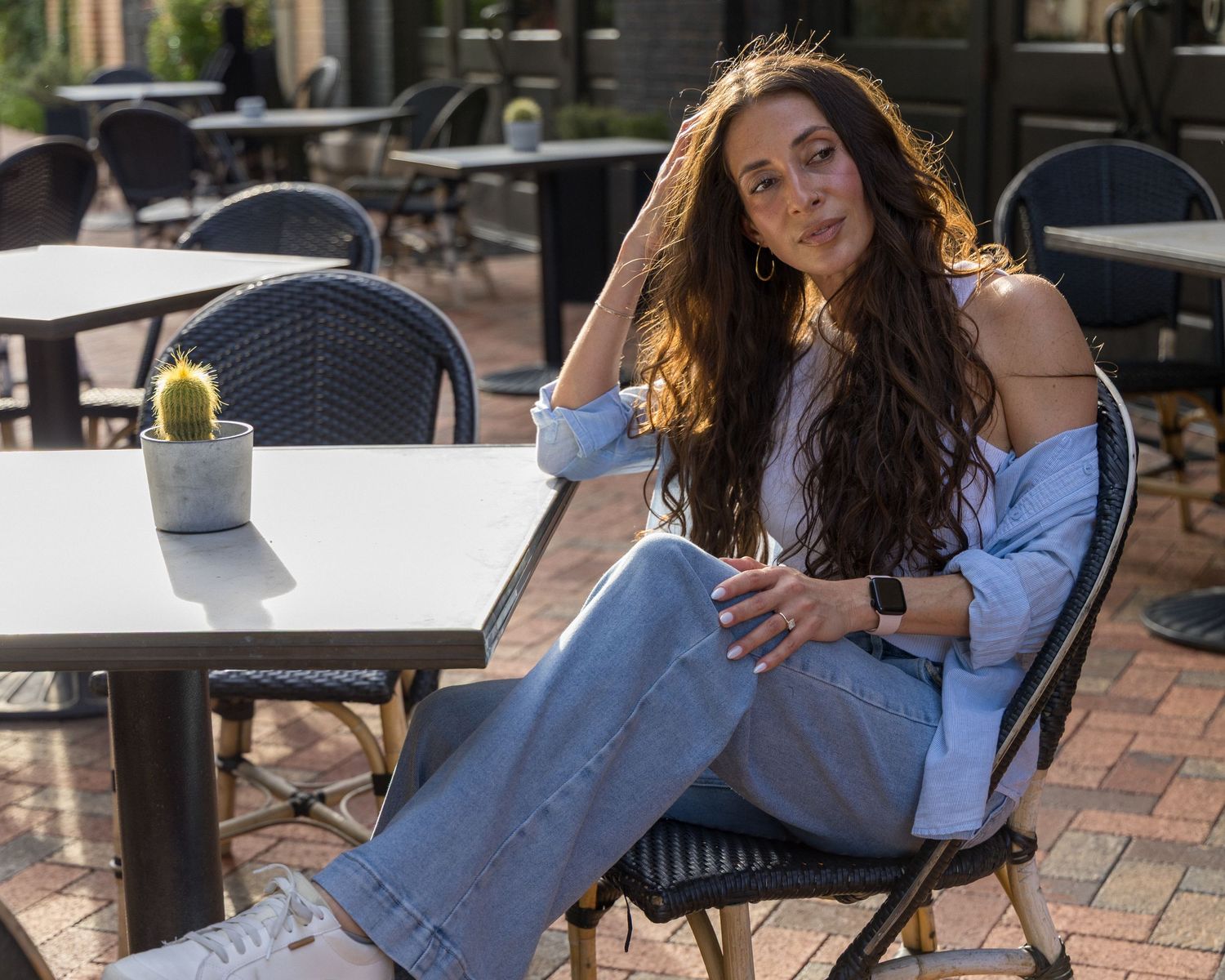 Woman with long wavy hair sits casually at outdoor patio table wearing blue shirt and wide-leg jeans.