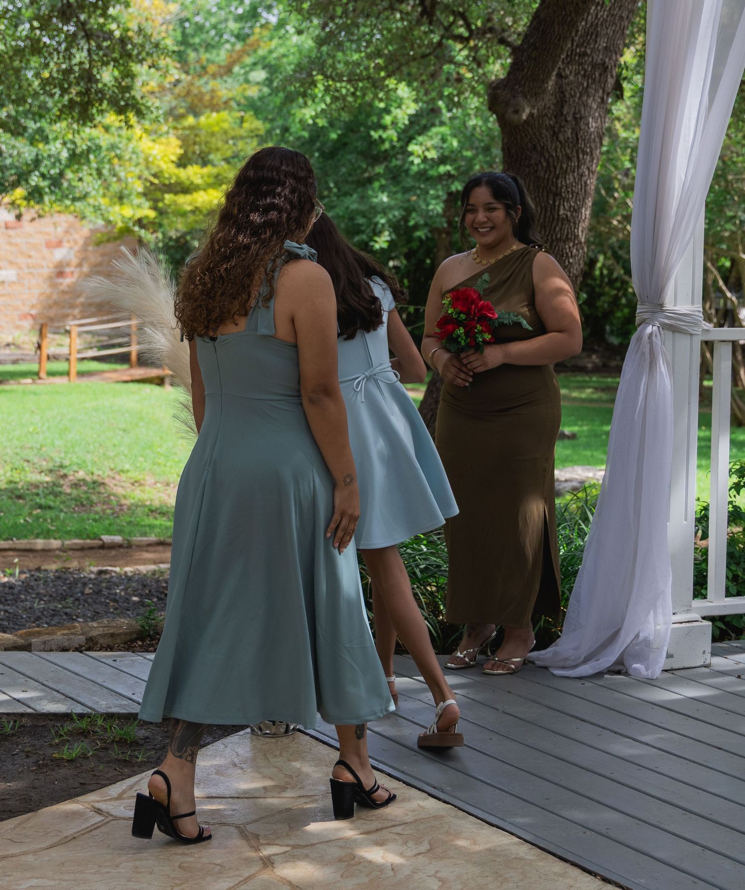 Bridesmaids in sage green dresses walk toward floral decorated outdoor wedding ceremony pergola.