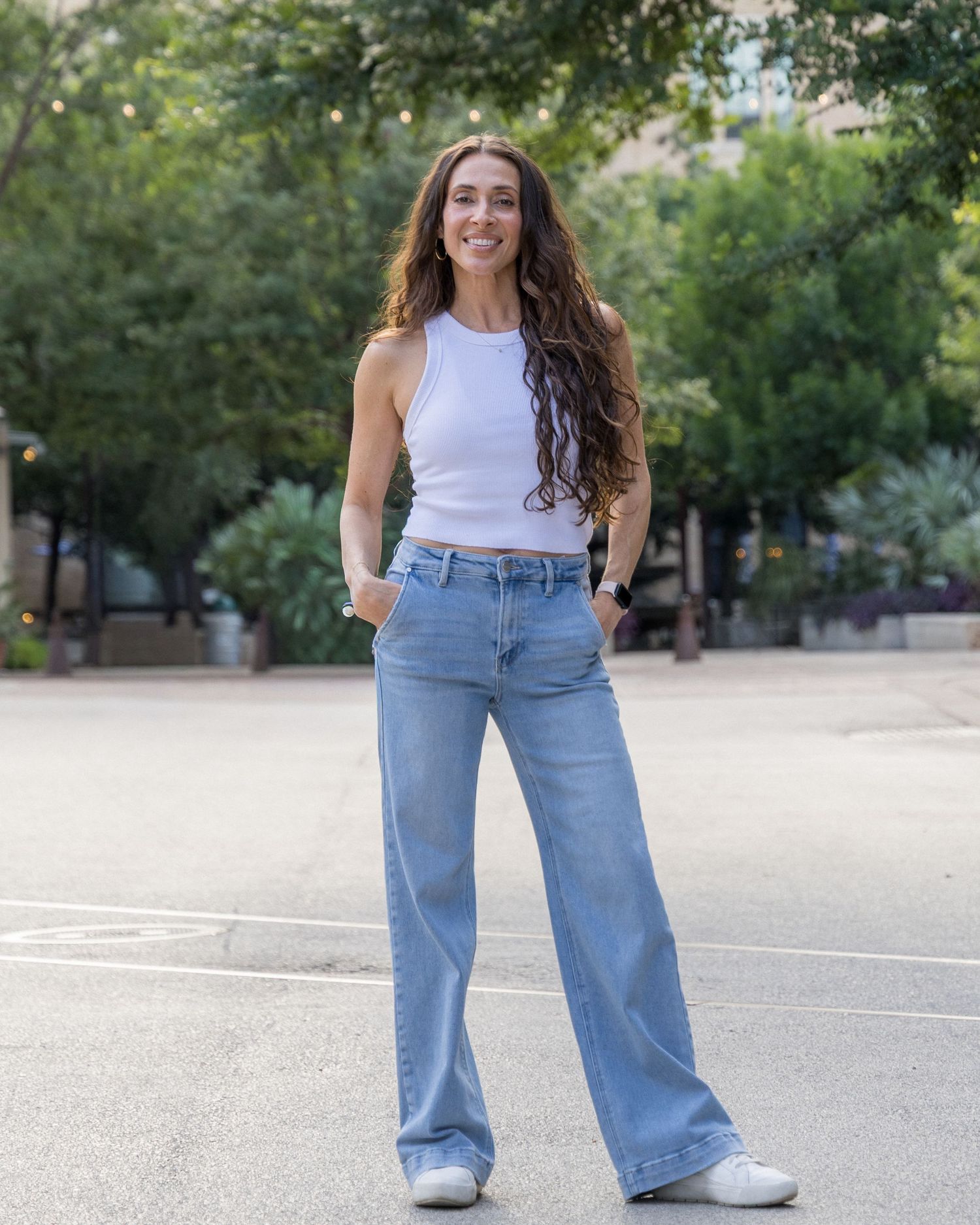 Woman in white tank top and wide-leg jeans stands confidently in urban plaza.