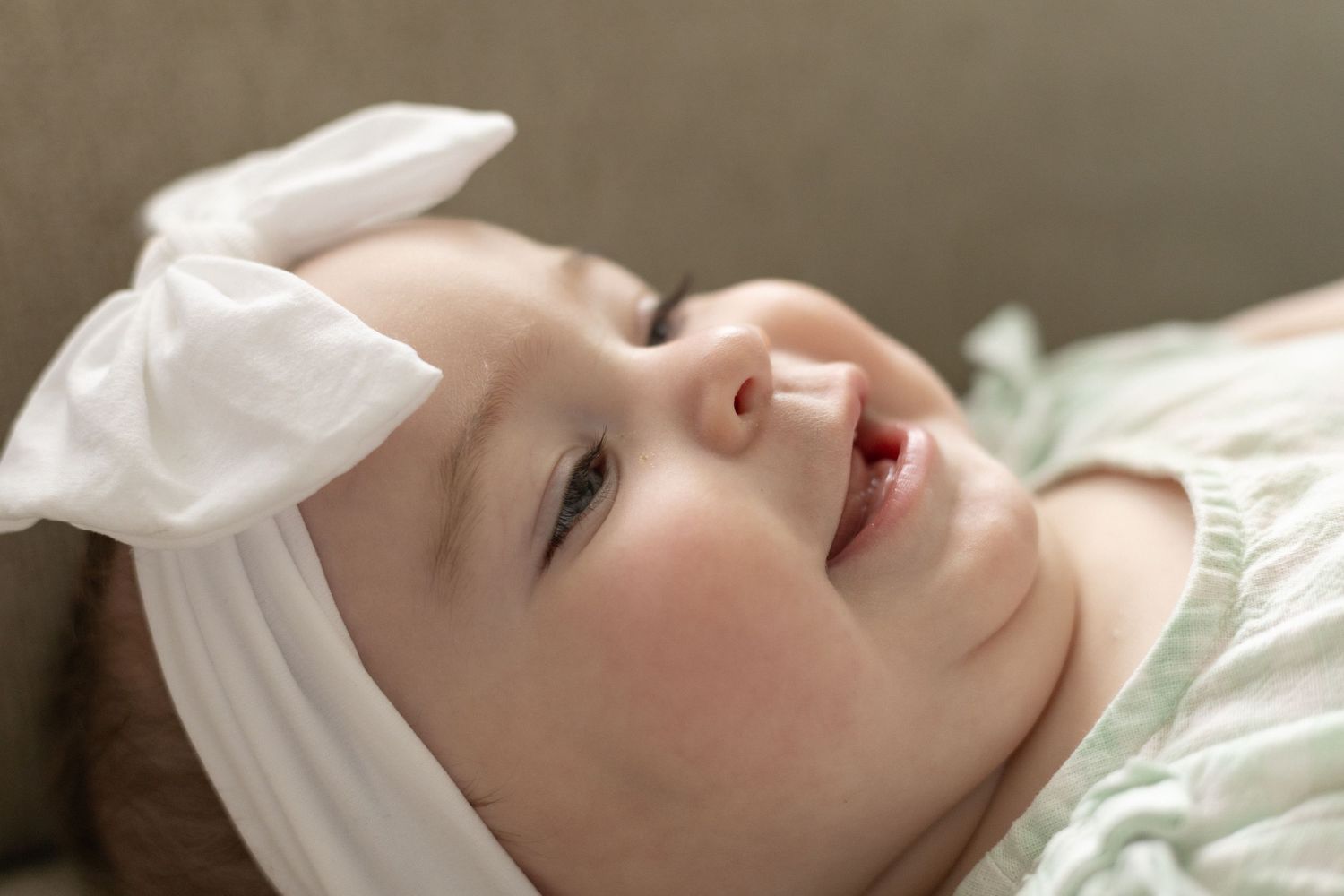 Adorable baby girl with white bow headband lying on sofa, looking up with bright eyes.