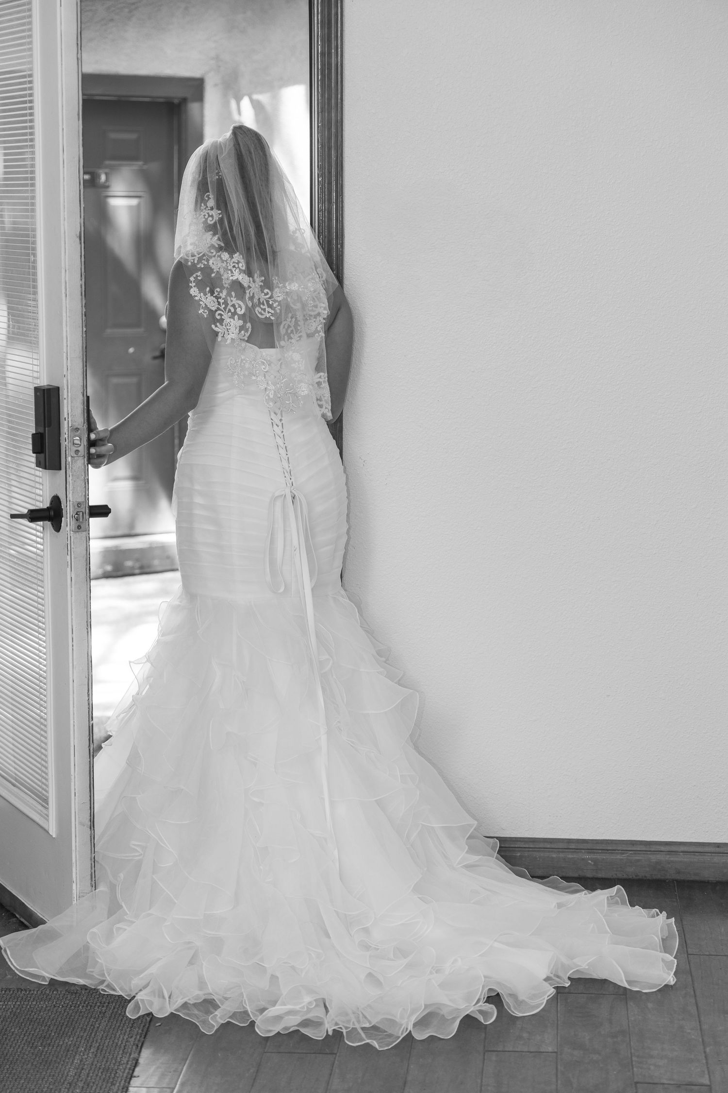 Black and white photo of a bride in a mermaid wedding gown with veil standing in a doorway.