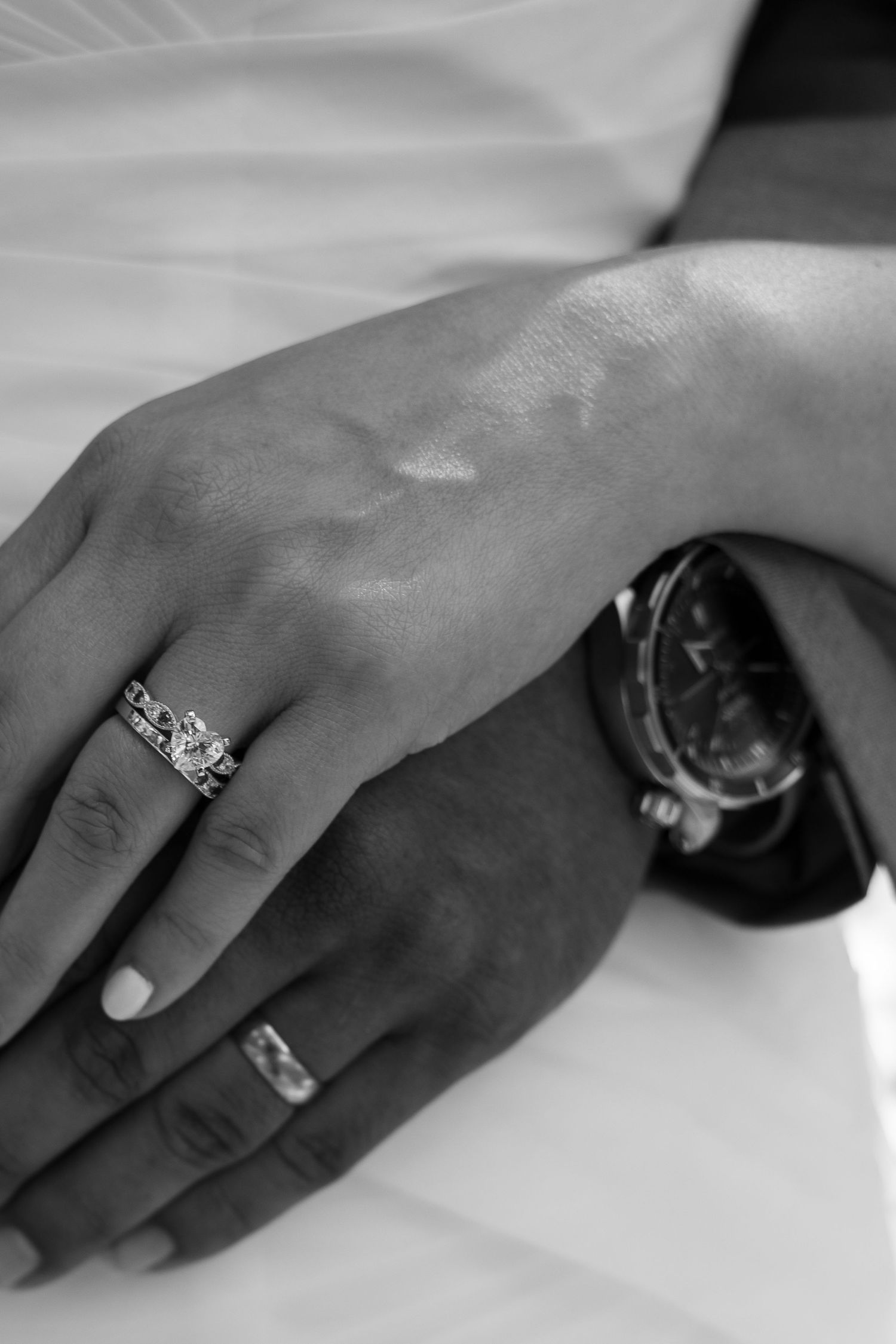 Black and white wedding photo of bride and groom's hands with engagement ring set and groom's watch and band.