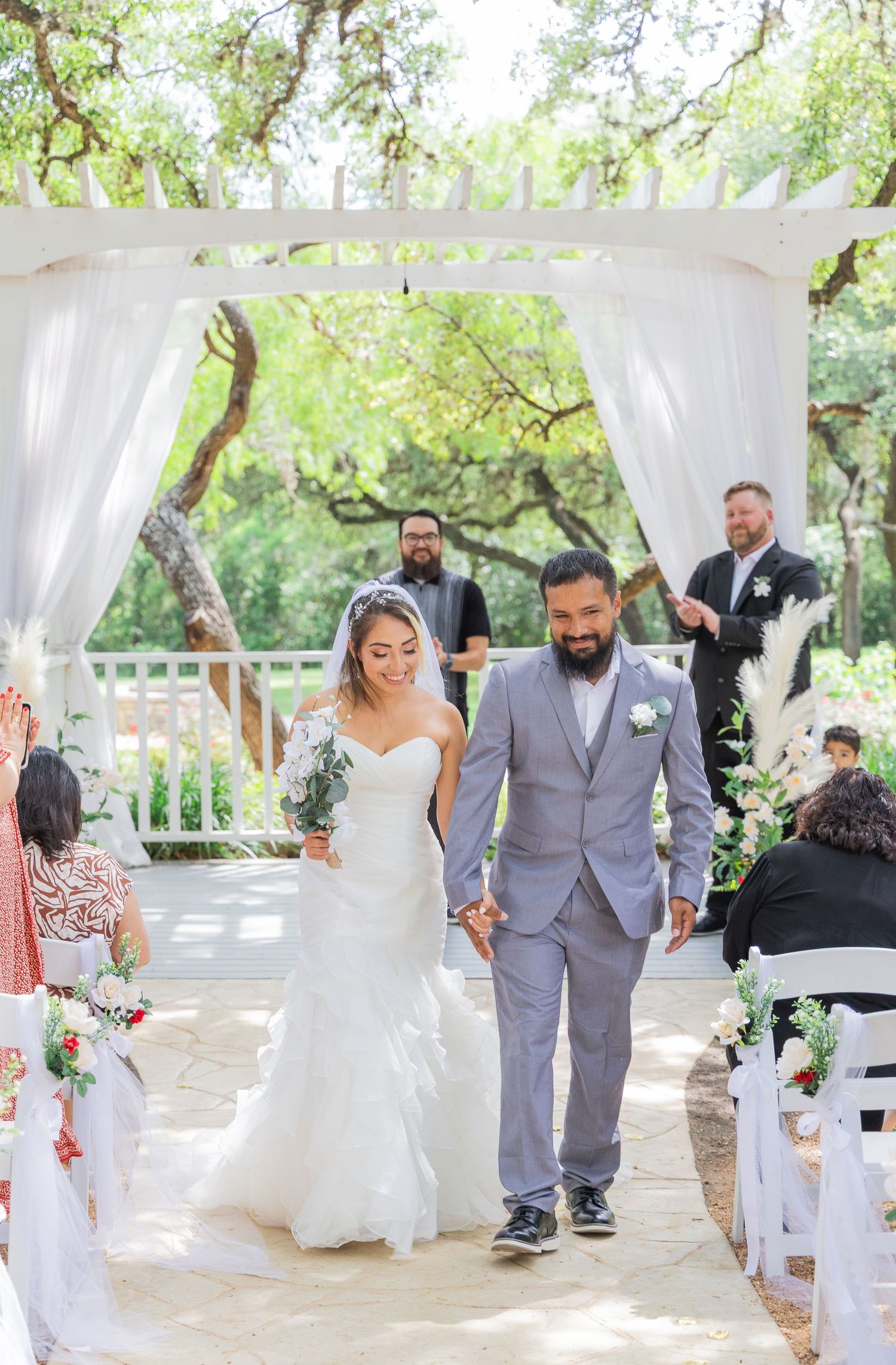 Newlyweds walk joyfully down aisle past seated guests after outdoor garden wedding ceremony.