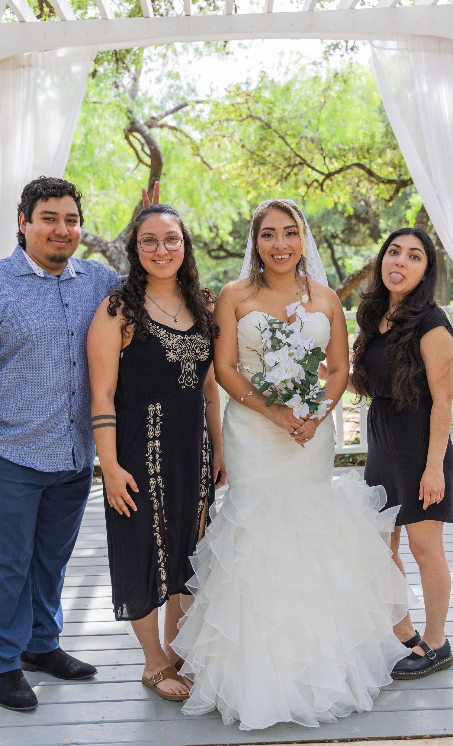 Bride in ruffled white gown holds white bouquet posing with three friends under outdoor wedding arch.