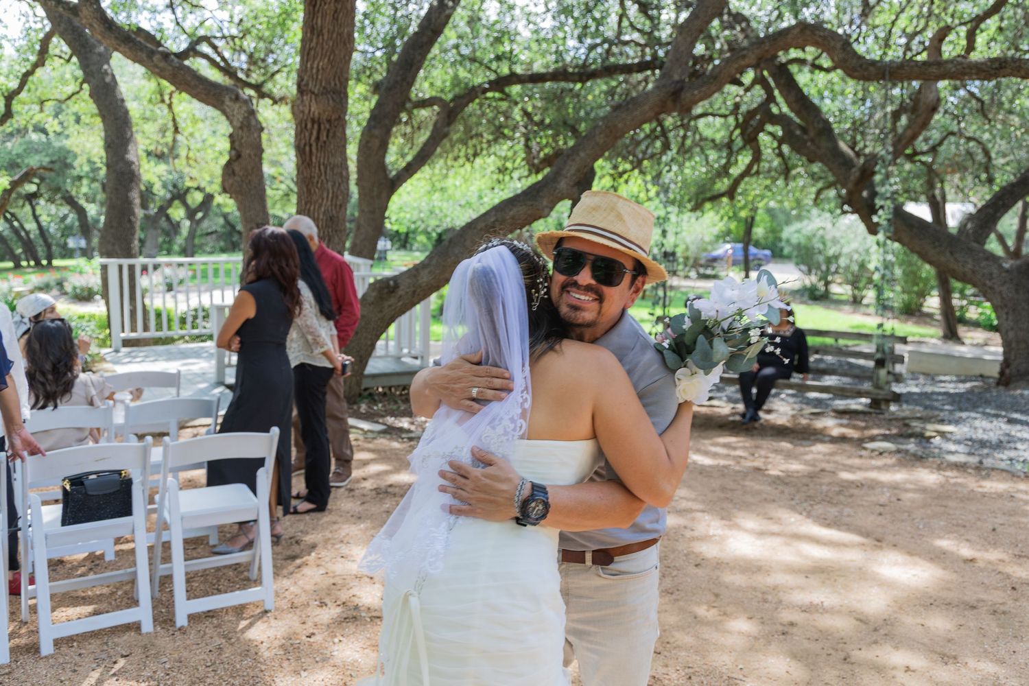 Groom in hat embracing bride in white gown near tree-lined outdoor wedding venue.