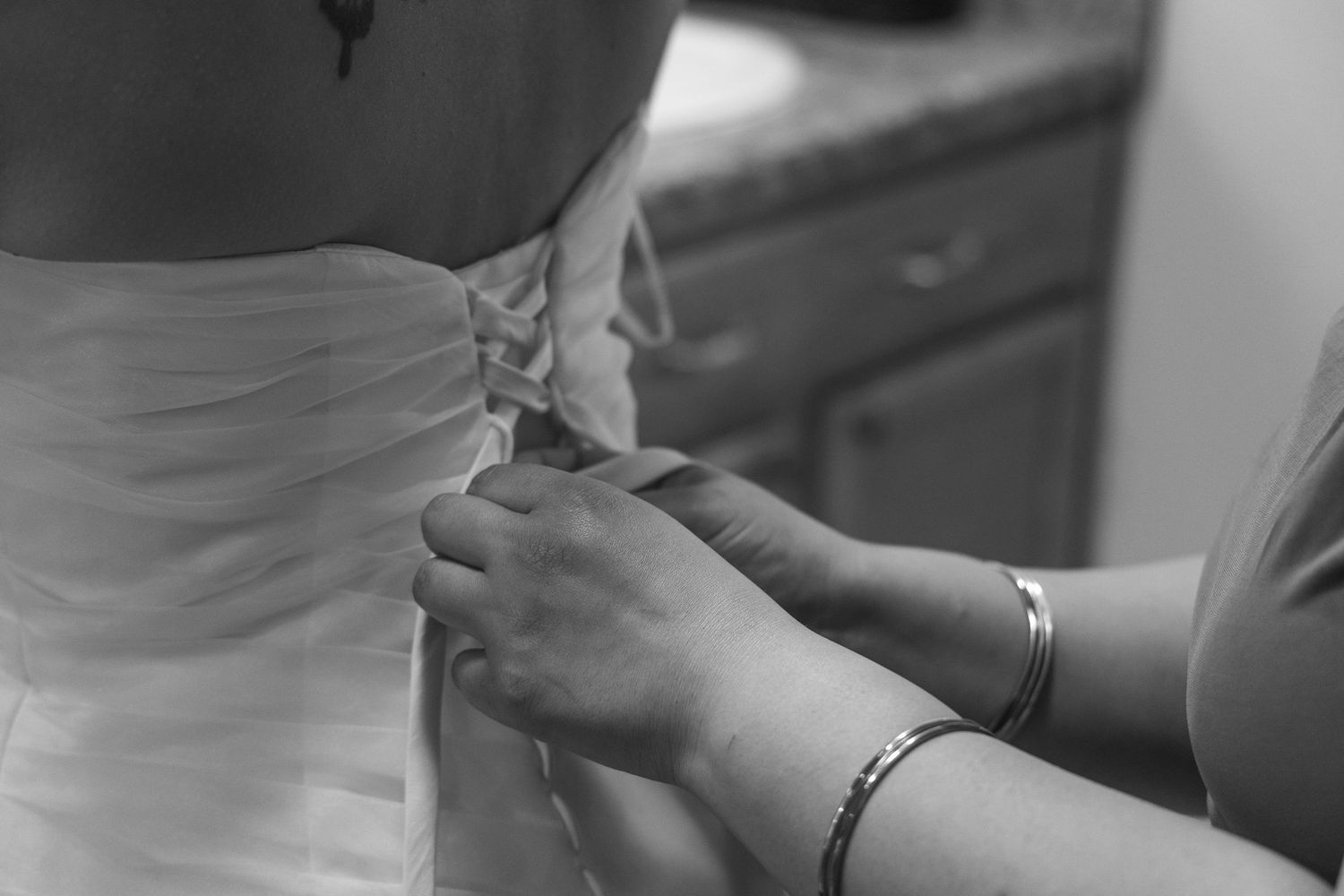 Black and white close-up of hands fastening buttons on the back of a white wedding dress.