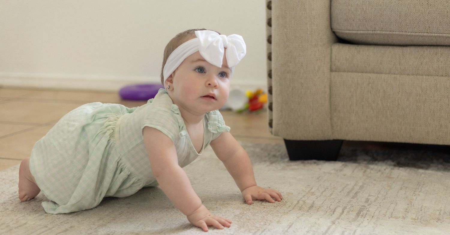 Baby girl in green romper and white bow headband crawling on carpet floor near beige armchair.