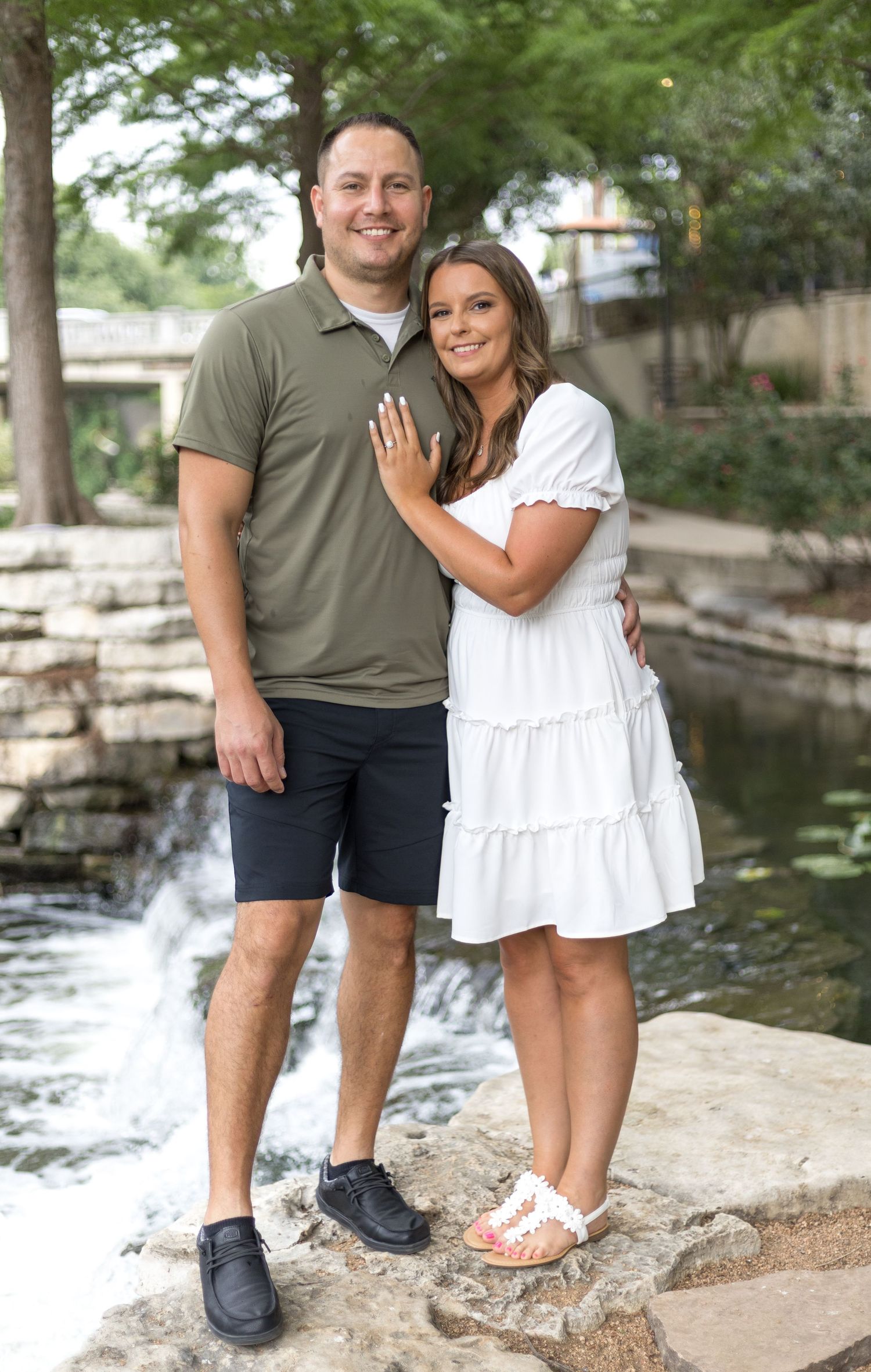 Smiling engaged couple posing together near a waterfall, woman showing off her engagement ring.