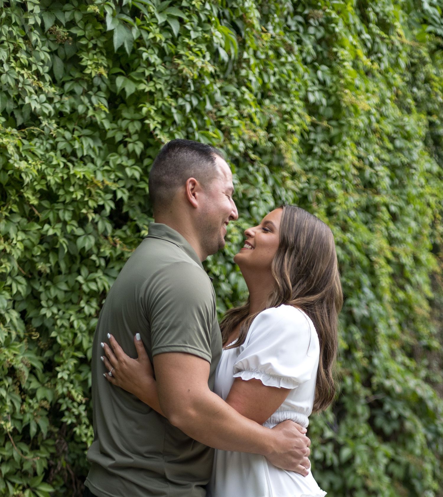 Engaged couple embracing and smiling at each other in front of a lush green ivy-covered wall.