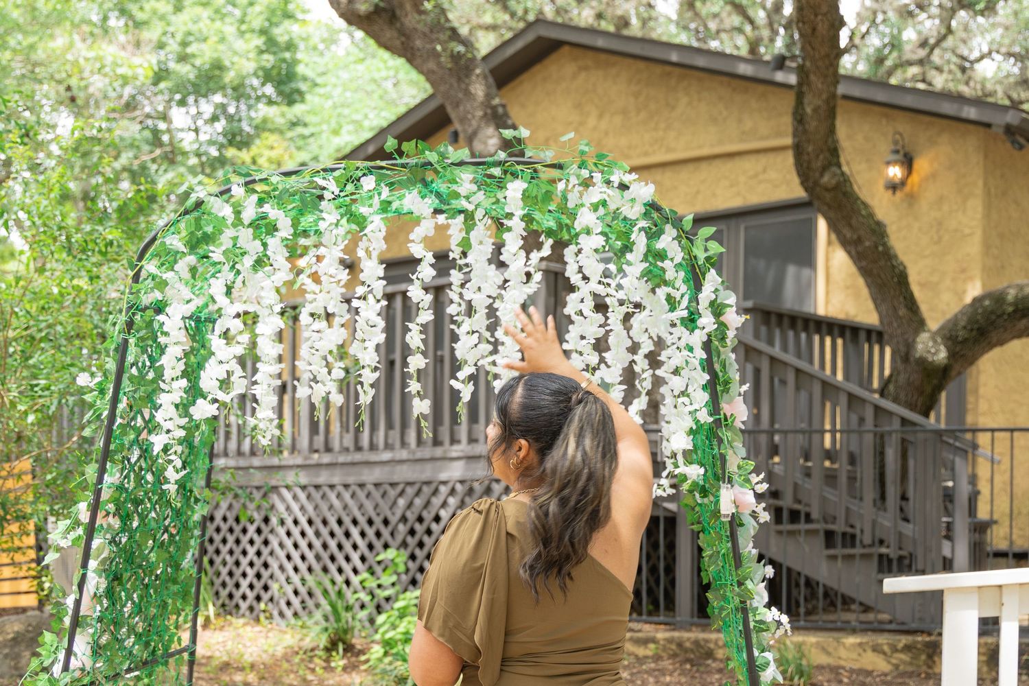 Woman decorating a floral wedding arch with white wisteria flowers outdoors near a wooden cabin.