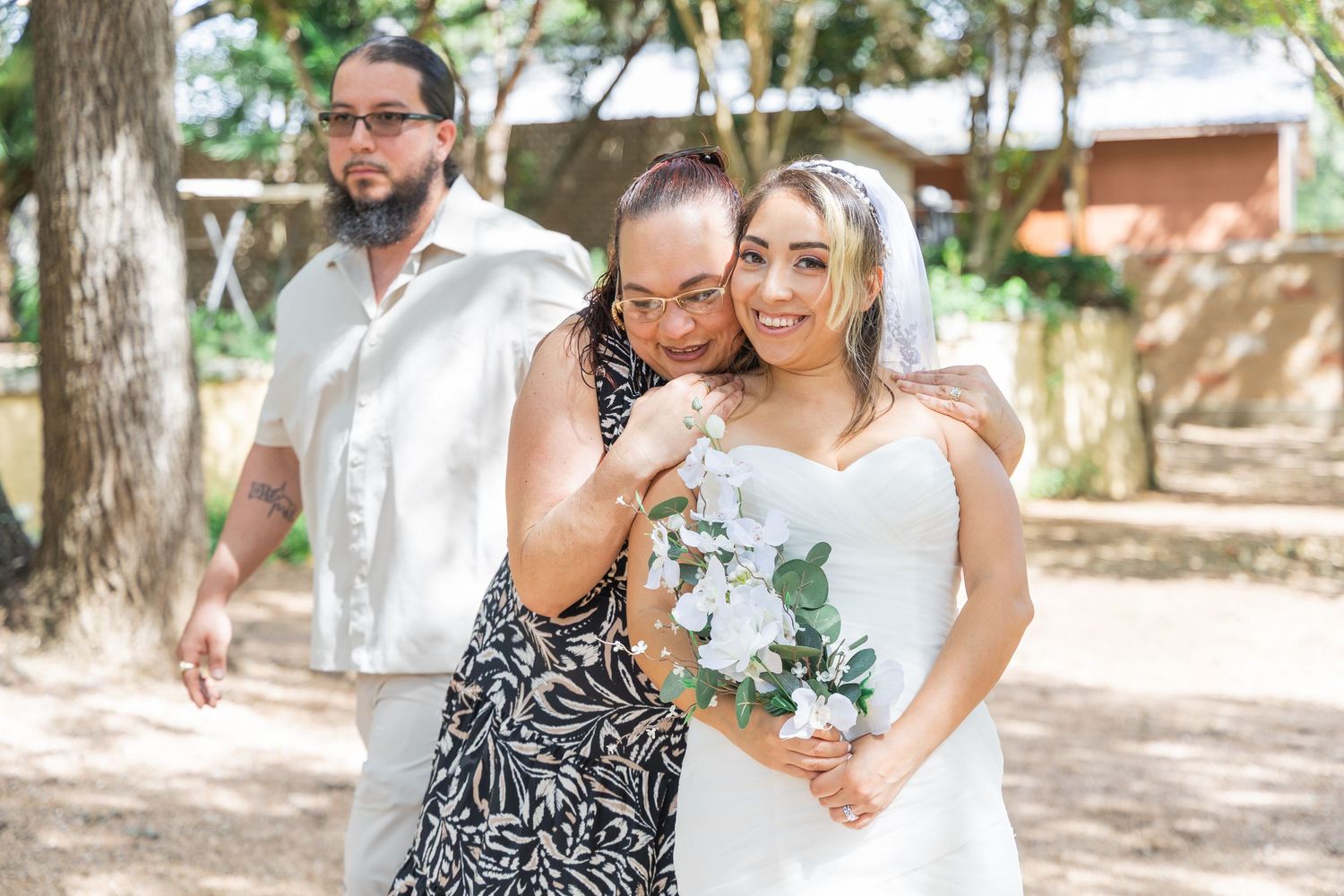 Bride in white dress holding bouquet posing with two family members outdoors.