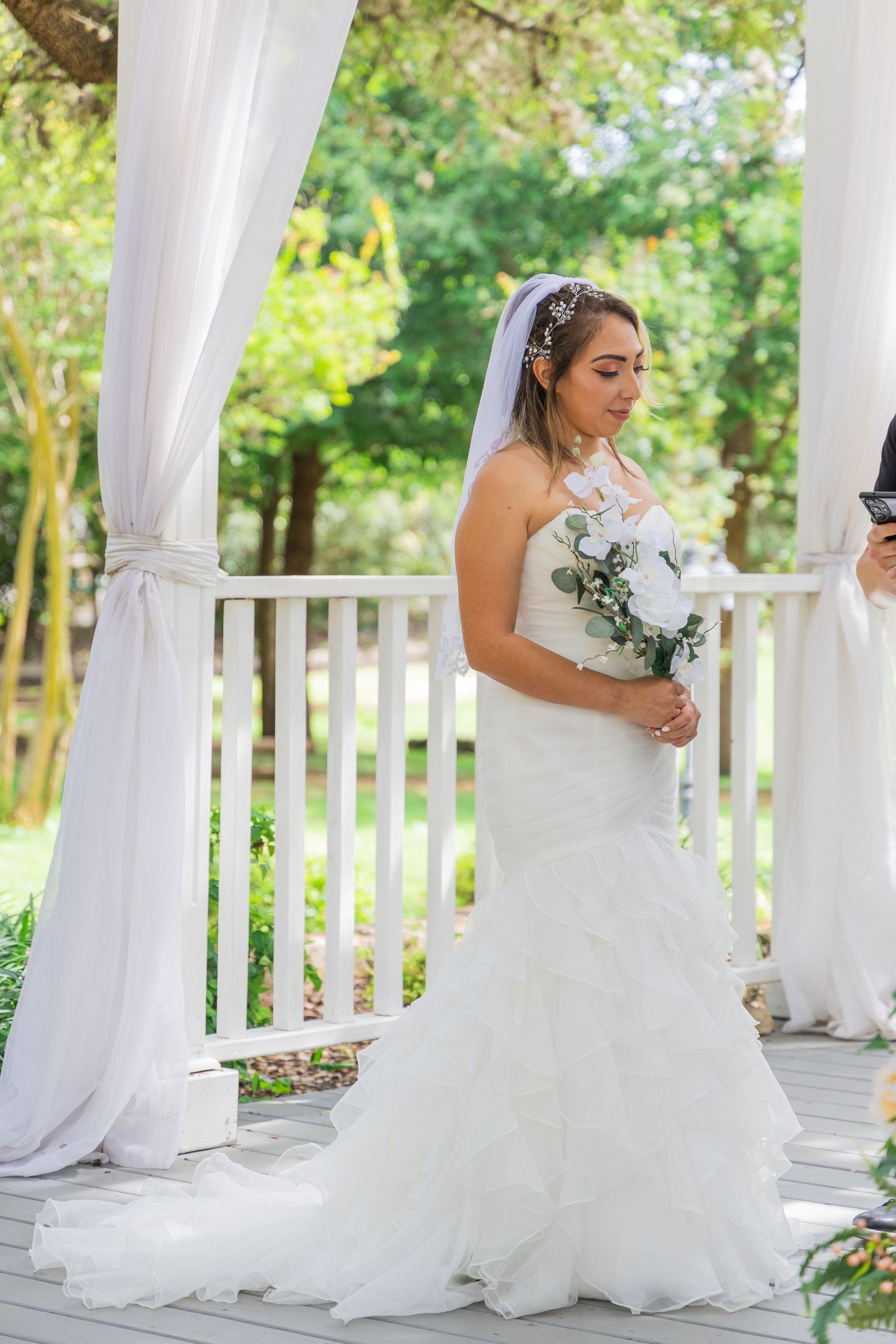Bride in strapless mermaid gown holding white bouquet stands under draped pergola at outdoor ceremony.