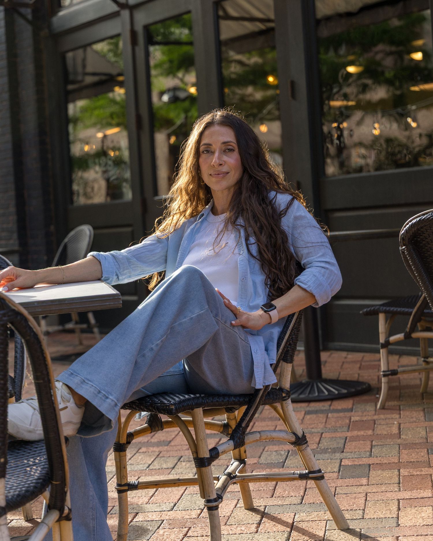 Woman with long dark hair sits elegantly in outdoor bistro chair wearing blue blazer and flare jeans.