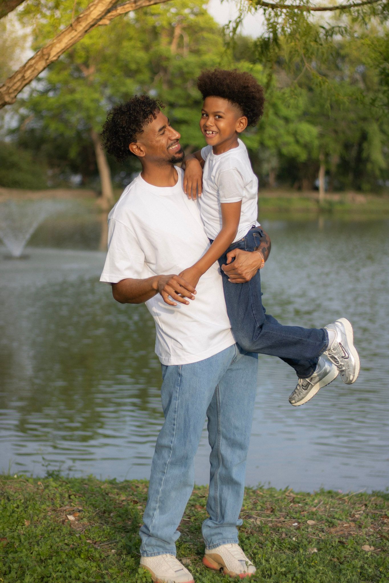 Happy father lifting his young son outdoors by a pond with a fountain and green trees.