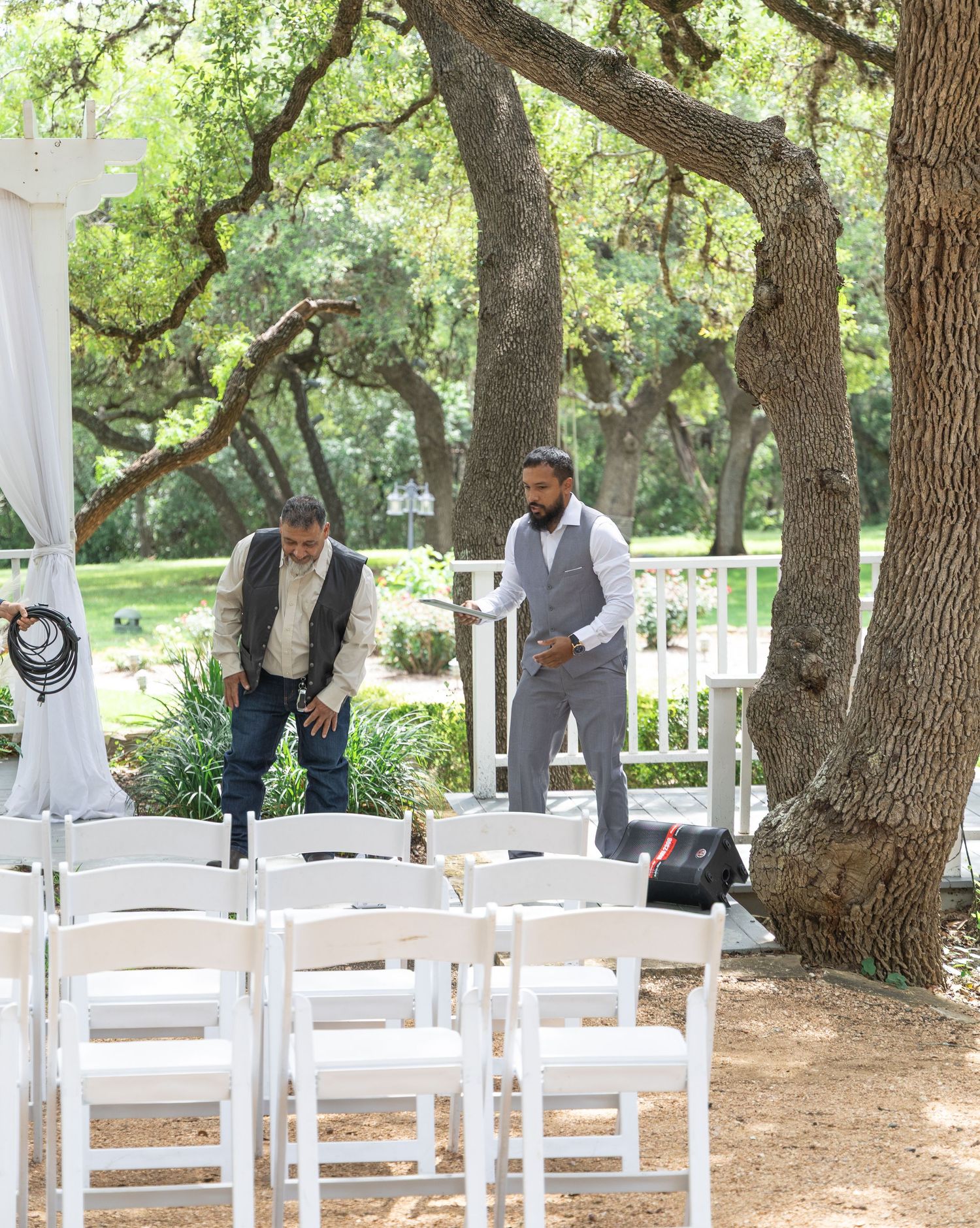 Two men in vests setting up white chairs for outdoor wedding ceremony near white pergola.