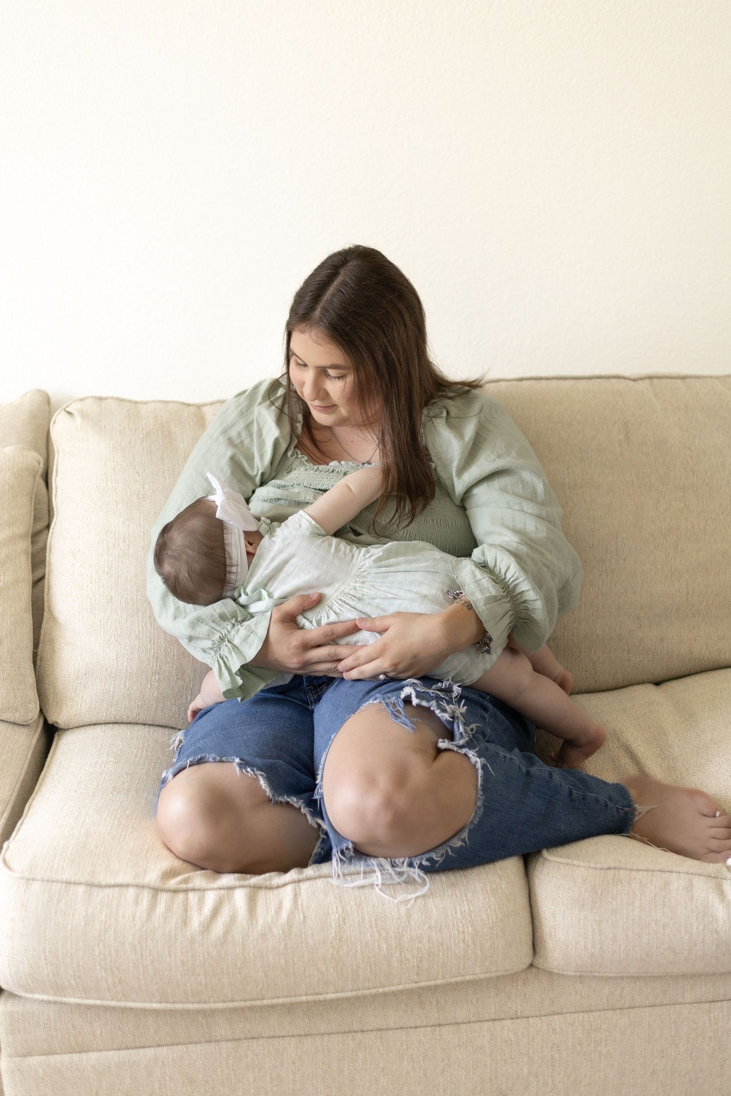 Young brunette mother in green blouse breastfeeding newborn baby while sitting on beige sofa.