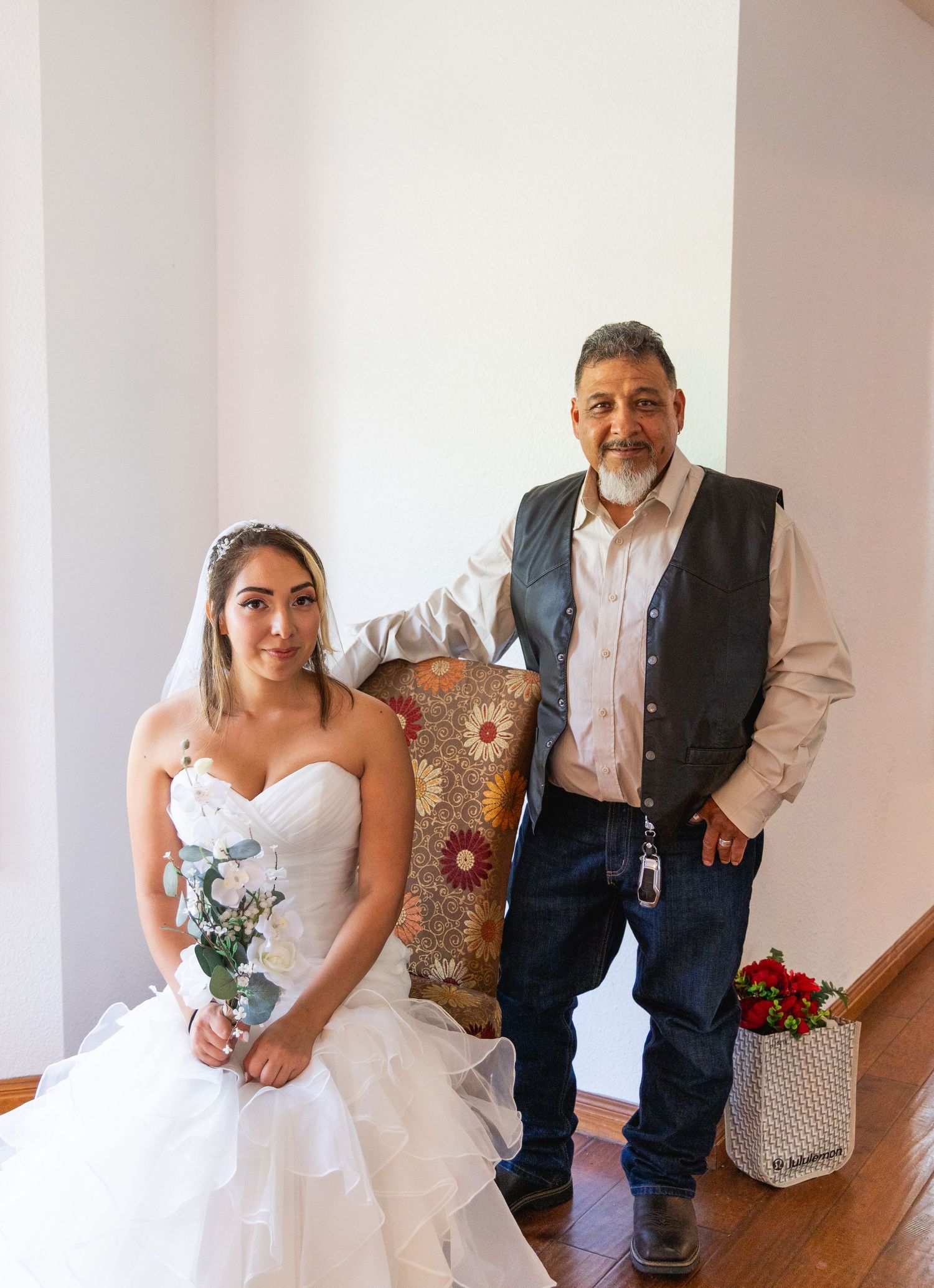 Bride in white strapless gown sitting beside older man in vest posing for wedding portrait indoors.