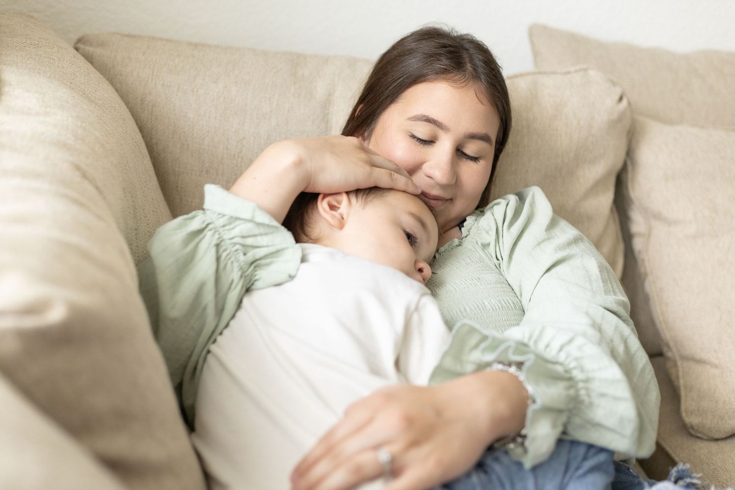 Loving mother in green top cuddling sleeping toddler boy on beige cushioned sofa at home.