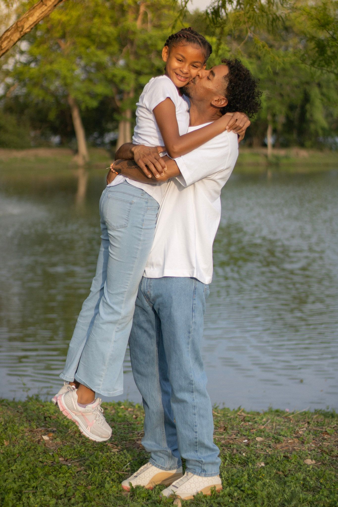 Father kissing his daughter on the cheek while lifting her near a sunlit park pond.