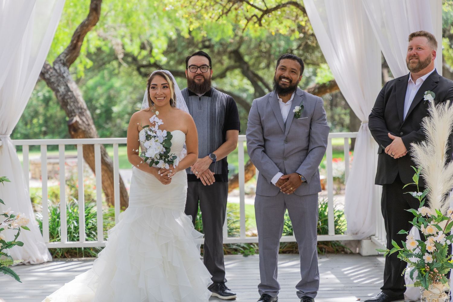Bride and groom stand at outdoor wedding altar with officiant and groomsman under white draped arbor.