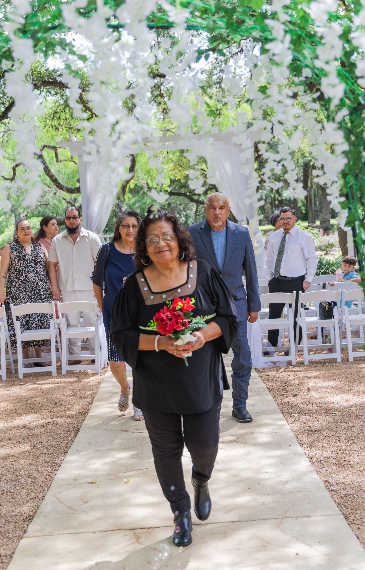 Woman in black outfit carries red flowers while leading guests down wedding aisle under floral arch.