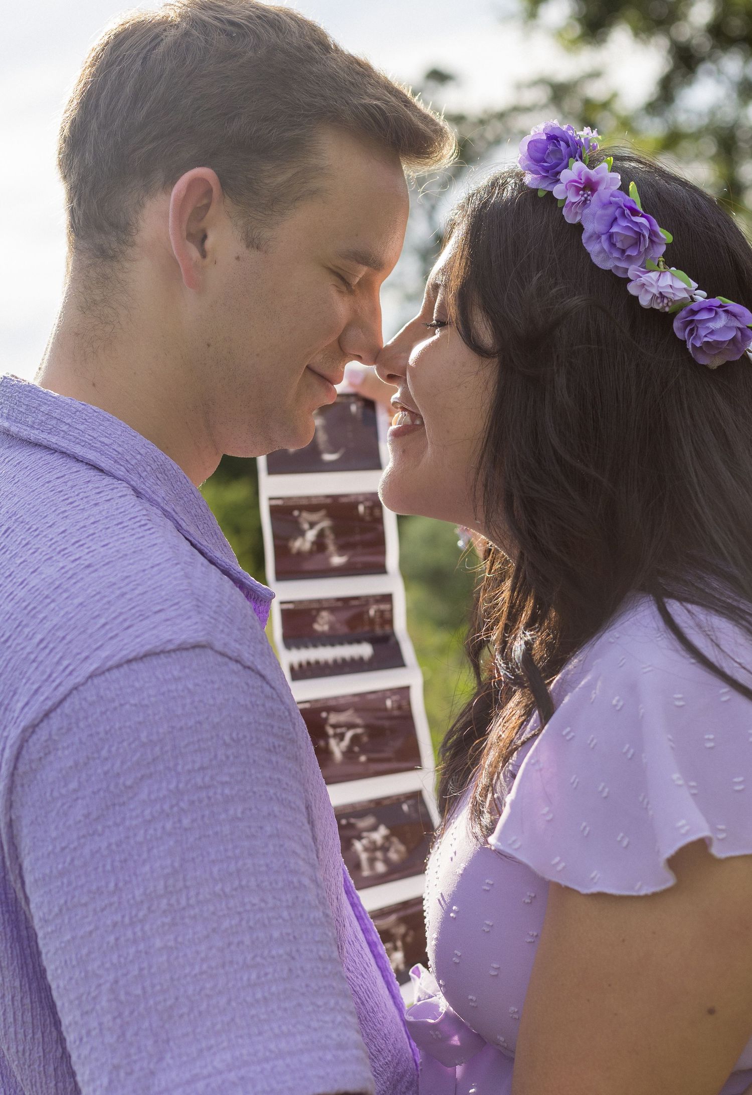 Expecting couple touches noses smiling while holding ultrasound photos at outdoor maternity photoshoot.