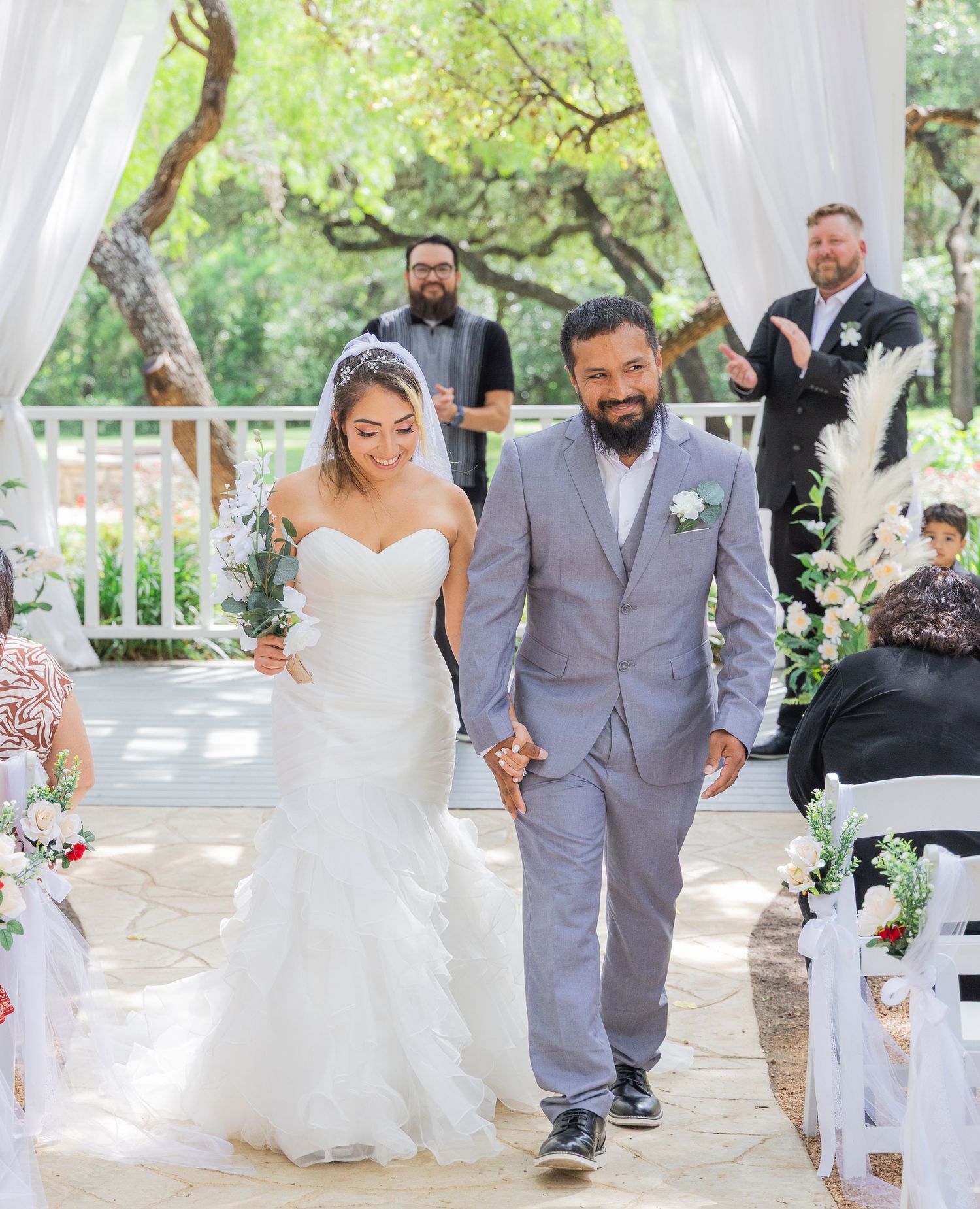 Bride and groom walk down the aisle hand in hand at an outdoor wedding ceremony under a white draped arch.