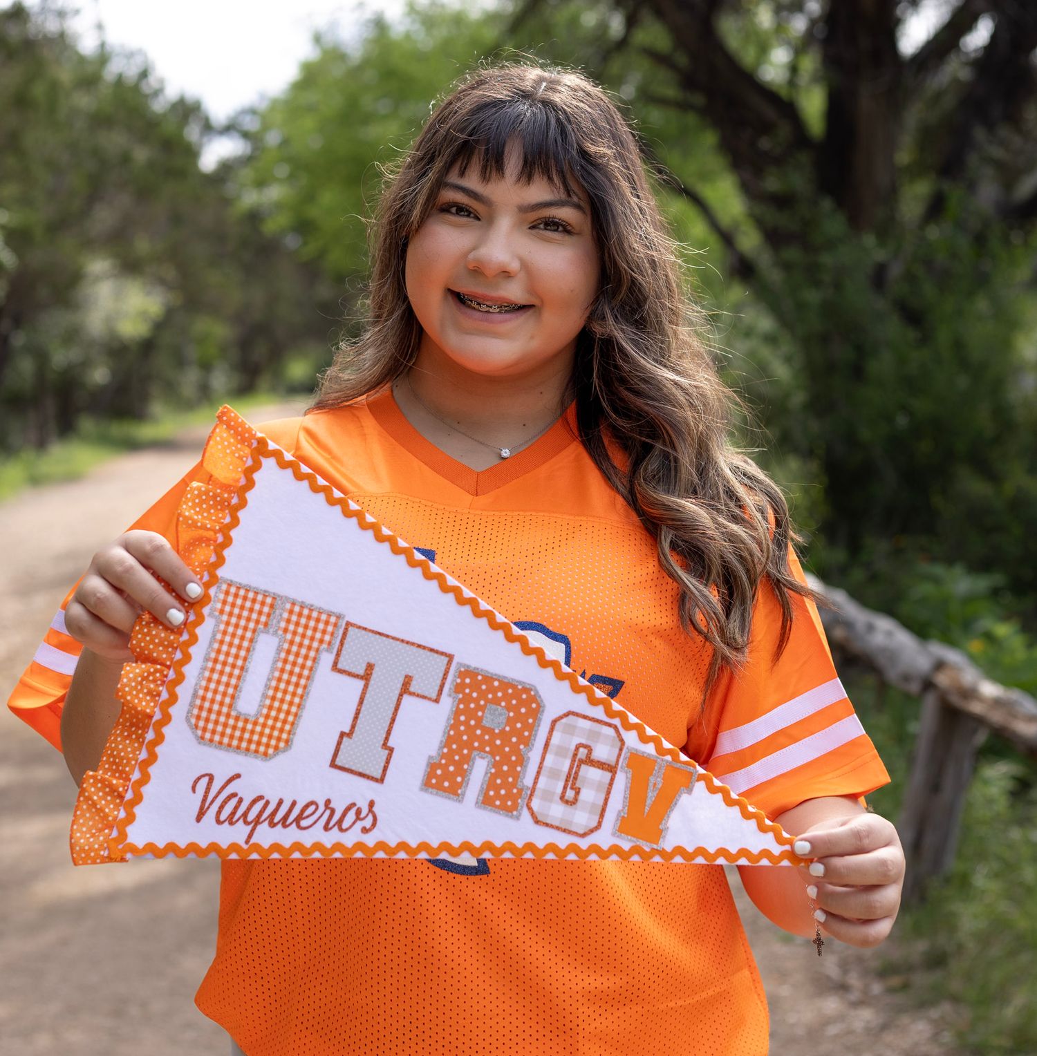 Smiling young woman holds UTRGV Vaqueros pennant flag wearing orange jersey outdoors.