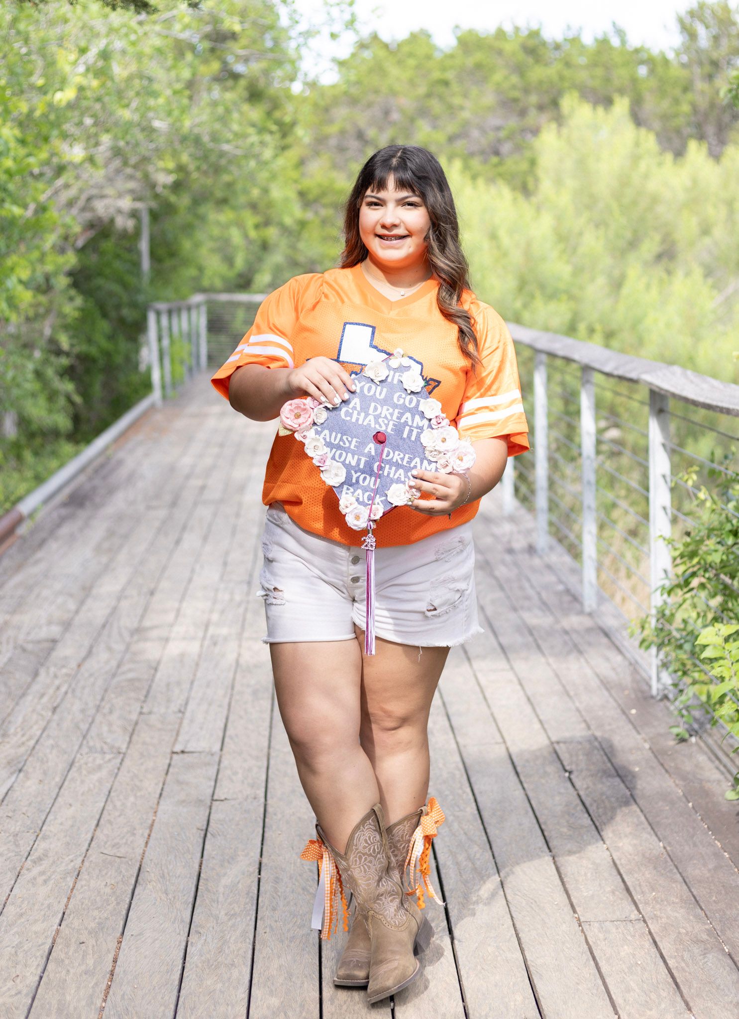 Smiling graduate in orange jersey holding Texas-shaped graduation cap on wooden bridge wearing cowboy boots.