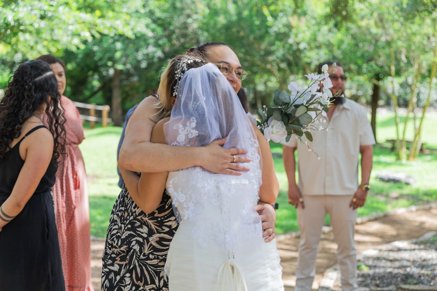 Bride in white gown and veil sharing an emotional hug with a guest outdoors.
