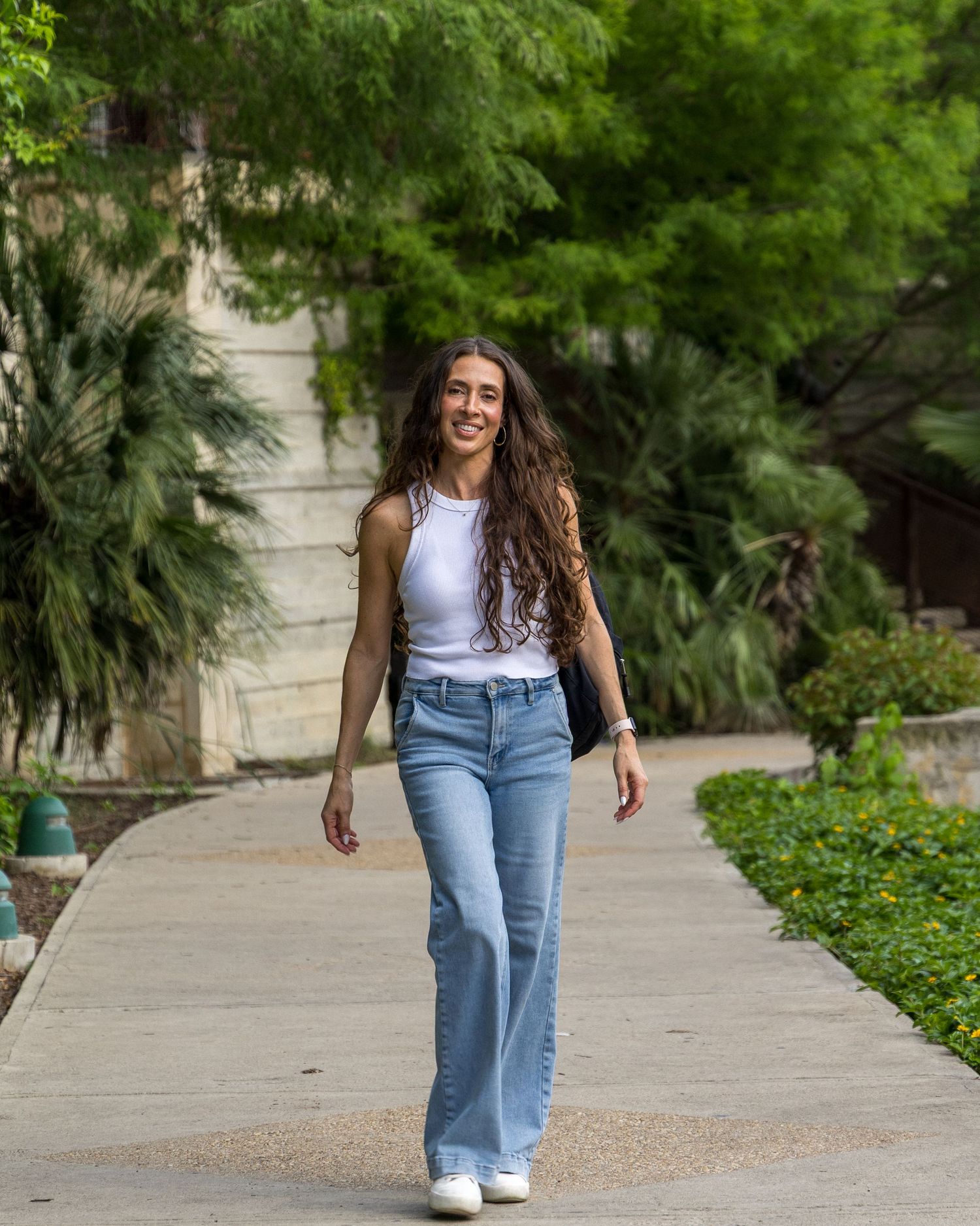 Young woman with long curly hair walks confidently in white tank top and light blue flare jeans on a sunny path.