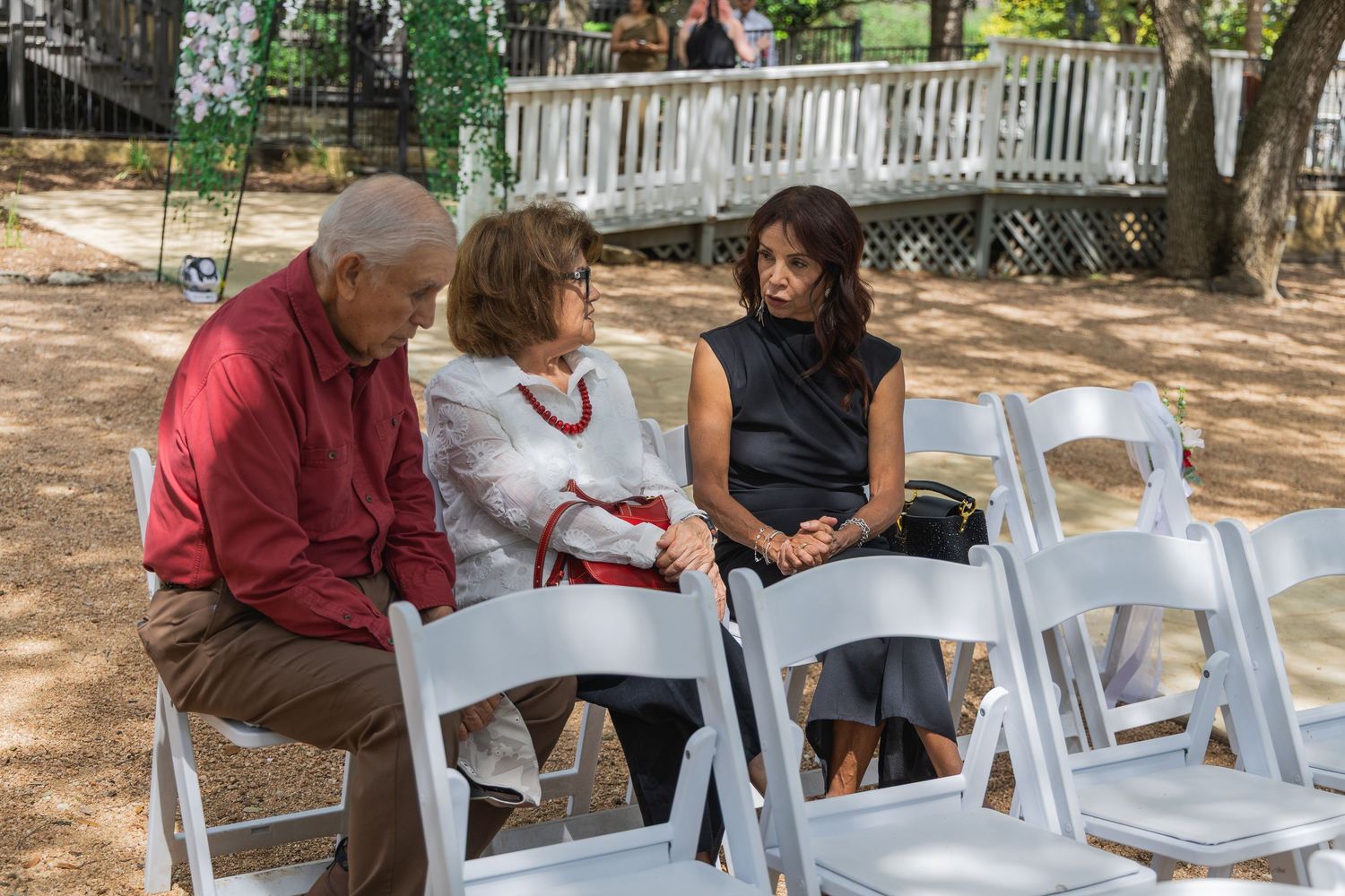 Three wedding guests in casual attire conversing while seated in white chairs at outdoor ceremony.