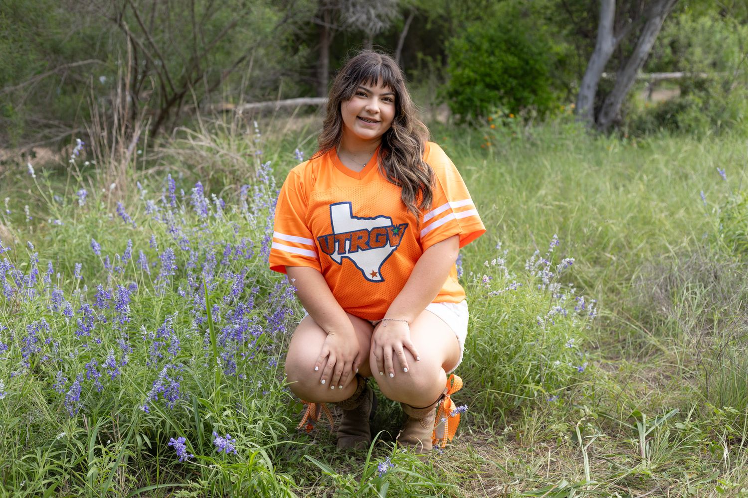 Young woman in orange UTRGV jersey crouches among purple wildflowers in field.