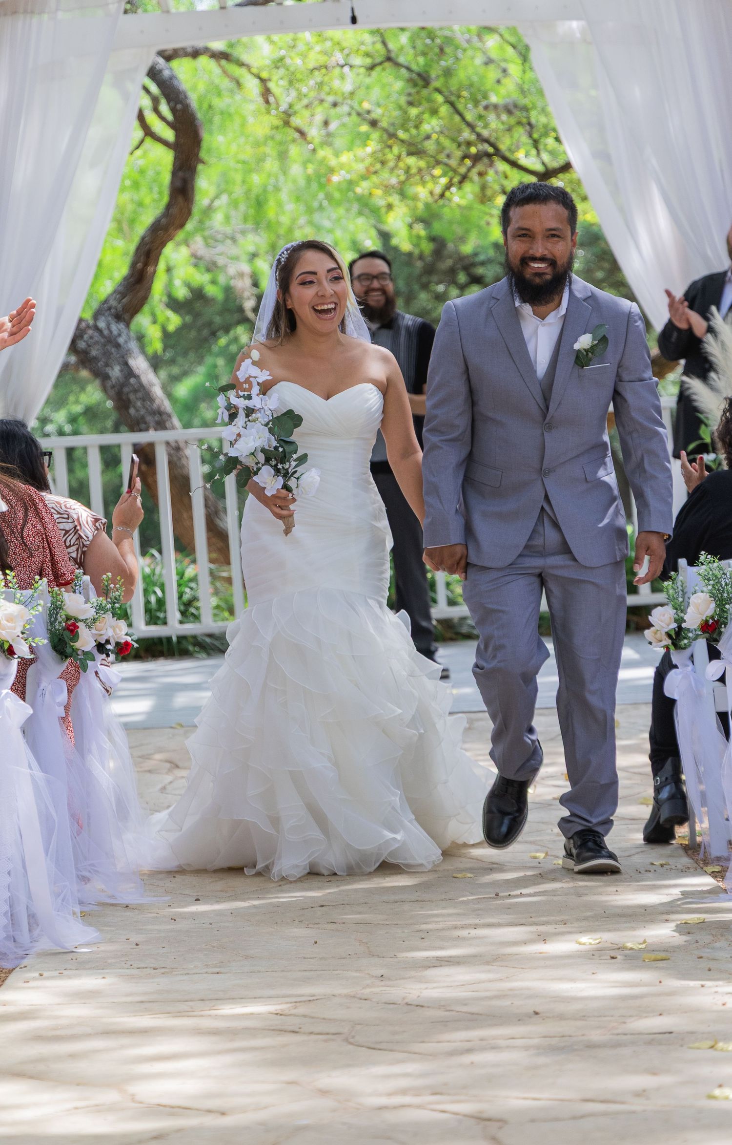 Joyful newlyweds walk down aisle after outdoor wedding ceremony surrounded by cheering guests.