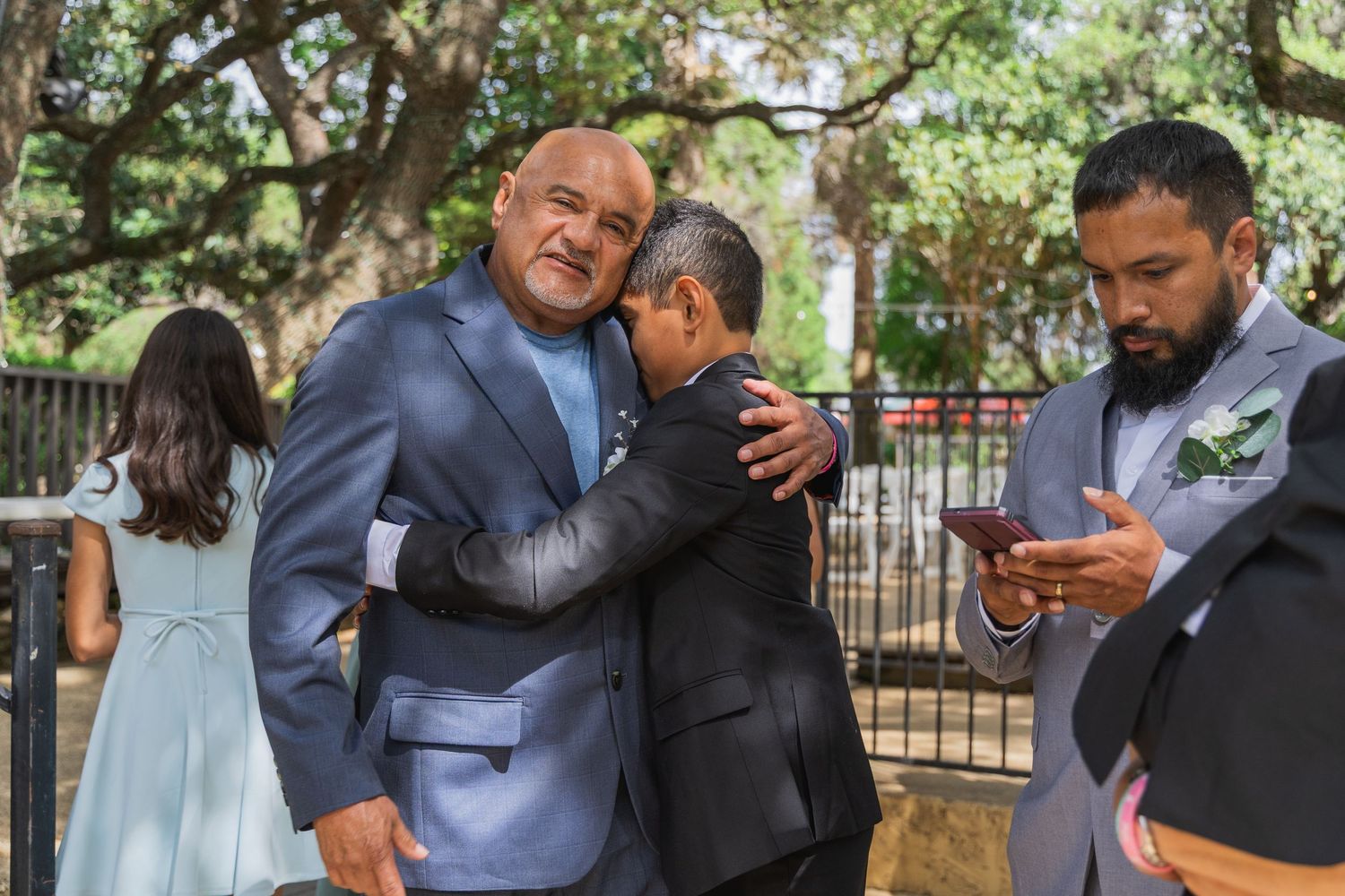 Two men share an emotional hug at outdoor wedding while another guest checks phone nearby.