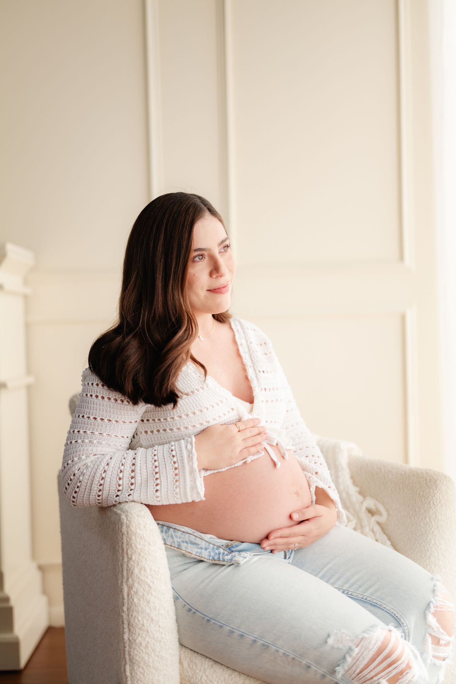 Pregnant woman in white crochet cardigan and jeans sits in armchair, gently cradling her baby bump.